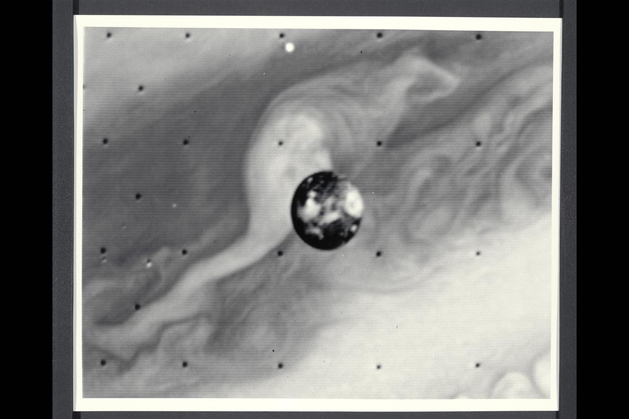 Range : 7 million kilometers (4.3 million miles) Io is Jupiter's innermost of the four Galilean satellites.  Photo taken at 2:00 AM through an ultraviolet filter. The photo's background is part of Jupiter's disk.  North is at the top and the central longitude of Io is 180 degrees.  Io shows a contrasting surface with dark polar areas and many light and dark regions around the equator.  This resolution of about 100 miles/160 kilometers, no topographic features, like craters, can be seen.  The brighter regions may be areas containing sulfur and various salts, making Io very reflective(six times brighter thanb Earth's Moon).  Io is about the same size and density as our Moon, but has followed a different evolutionary path, influenced by its closeness to Jupiter and the intense bombardment it receives from the Jovian radiation belts of energetic charged particles.