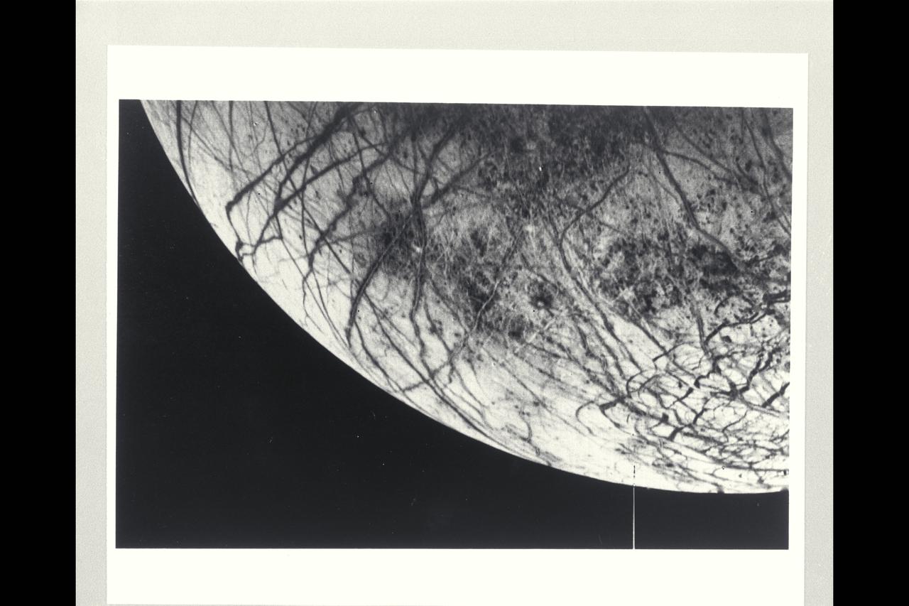 P-21760 BW This color image of the Jovian moon Europa, which is the size of our moon, is thought to have a crust of ice perhaps 100 kilometers thick which overlies the silicate crust. The complex array of streaks indicate that the crust has been fractured and filled by materials from the interior. The lack of relief, any visible mountains or craters, on its bright limb is consistent with a thick ice crust. In contrast to its icy neighbors, Ganymede and Callisto, Europa has very few impact craters. One possible candidate is the small feature near the center of this image with radiating rays and a bright circular interior. The relative absence of features and low topography suggests the crust is young and warm a few kilometers below the surface. The tidal heating process suggested for Io also may be heating Europa's interior at a lower rate.