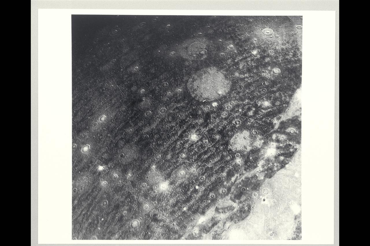 This color reconstruction of part of the northern hemisphere of Ganymede was made from pictures taken at a range of 313,000 km (194,000 mi.). The scene is approx. 1,300 km (806 mi.) across. It shows part of dark, densely cratered block which is bound on the south by lighter, and less cratered, grooved terrain. The dark blocks are believed to be the oldest parts of Ganymede's surface. Numerous craters are visible, many with central peaks. The large bright circular features have little relief and are probably the remnants of old, large craters that have been annealed by flow of the icy near-surface material. The closely-spaced arcuate, linear features are probably analogous to similar features on Ganymede which surround a large impact basin. The linear features here may indicate the former presence of a large impact basin to the southwest.