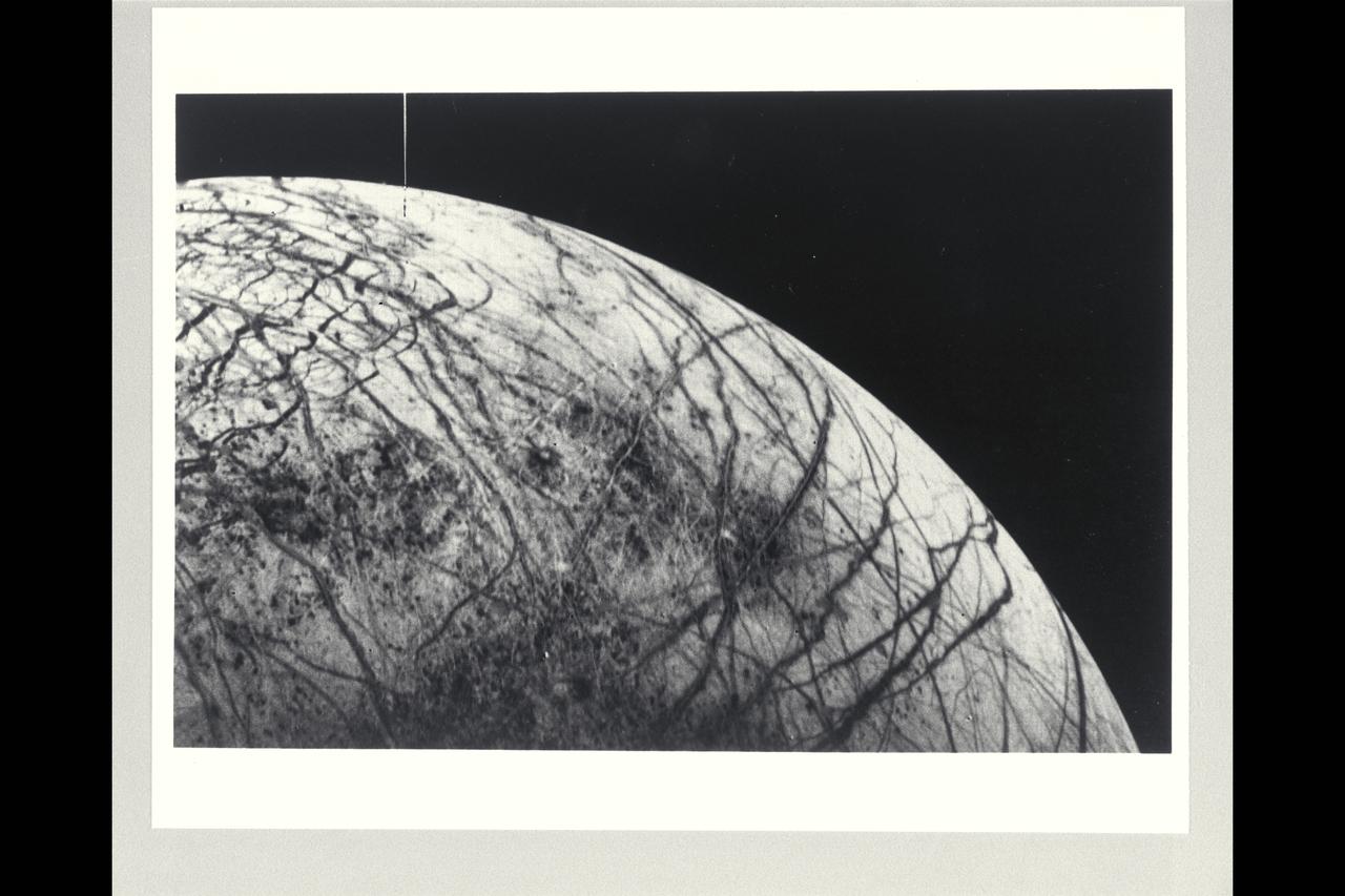 Range :  241,000km (150,600 mi.). This black and white image of Europa, smallest of Jupiter's four Galilean satellites, was acquired by Voyager 2.  Europa, the brightest of the Galiliean satellites, has a density slightly less than Io, suggesting it has a substantial quantity of water.  Scientists previously speculated that the water must have cooled from the interior and formed a mantle of ice perhaps 100 km thick.  The complex patterns on its surface suggest that the icy surface was fractured, and that the cracks filled with dark material from below.  Very few impact craters are visible on the surface, suggesting that active processes on the surface are still modifying Europa.  The tectonic pattern seen on its surface differs drastically from the fault systems seen on Ganymede where pieces of the crust have moved relative to each other.  On Europa, the crust evidently fractures but the pieces remain in roughly their original position.