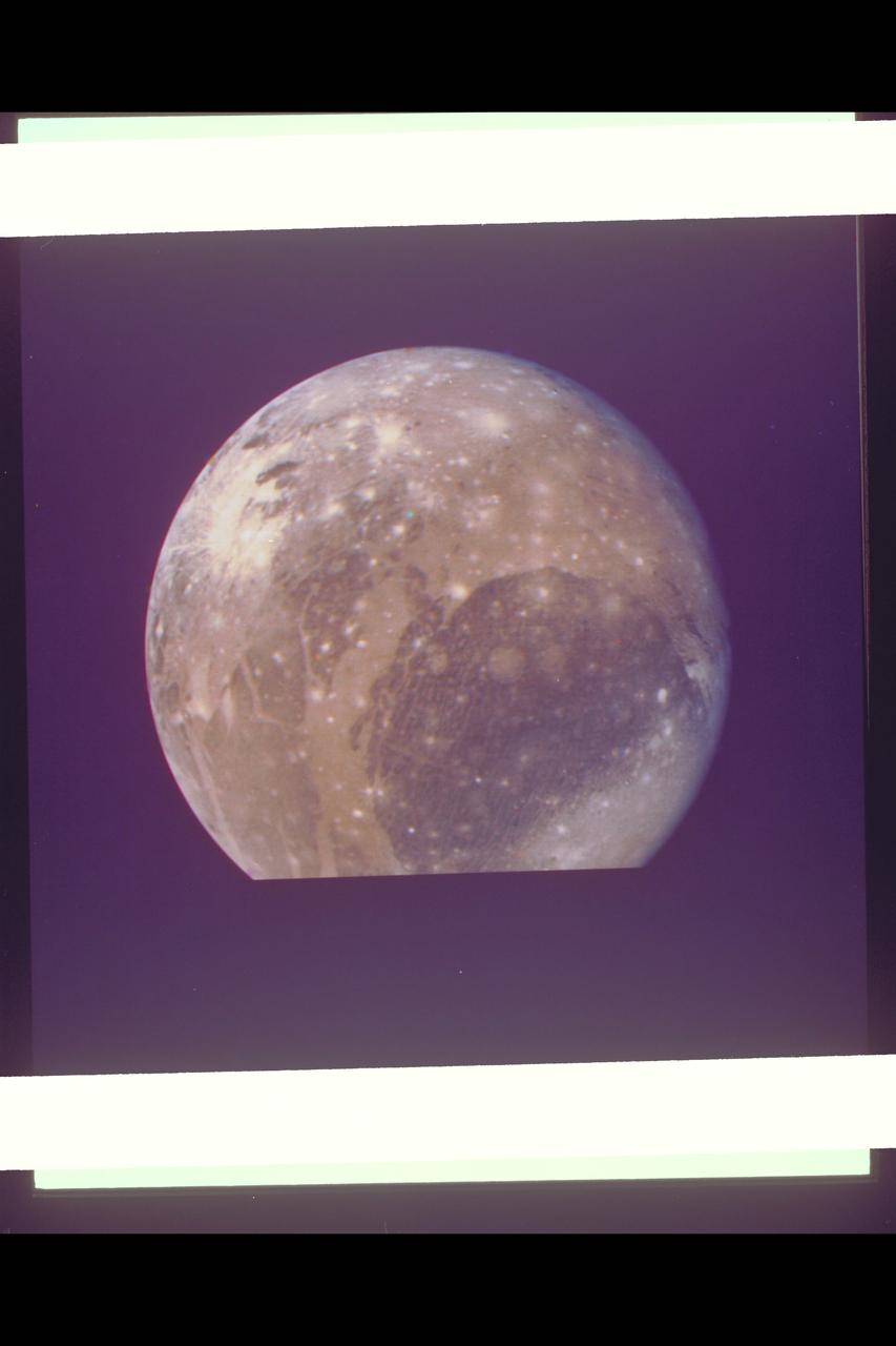 P-21751 C Range: 1.2 million kilometers This Voyager 2 color photo of Ganymede, the largest Galilean satellite, shows a large dark circular feature about 3200 kilometers in diameter with narrow closely-spaced light bands traversing its surface. The bright spots dotting the surface are relatively recent impact craters, while the lighter circular areas may be older impact areas. The light branching bands are ridged and grooved terrain first seen on Voyager 1 and are younger than the more heavily cratered dark regions. The nature of the brightish region covering the northern part of the dark circular fature is uncertain, but it may be some type of condensate. Most of the features seen on the surface of Ganymede are probably both internal and external responses of the very thick icy layer which comprises the crust of this satellite.