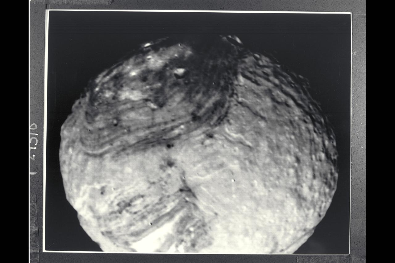 P-29510BW Range: 150,000 kilometers (90,000 miles) Nearly the full disk of Miranda can be seen in this clear-filter image taken by Voyager's narrow-angle camera.  At this resolution, about 2.7 km (1.7 mi), only two distinct terrain types are visible. One is a bright, rugged, heavily-cratered region seen along the bottom of the disk and extending to the top in a narrow band. The second terrain type is a topographically lower, darker, grooved terrain along the two upper sides in this image. The dark areas had been visible for several days before closest approach in lower-resolution views of the satellite. Scarps, or cliffs, visible along the limbs have a rugged relief of several kilometers in height.