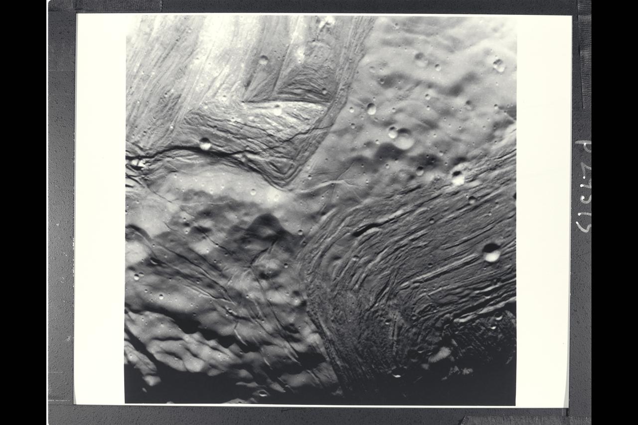 P-29515 BW Range: 42,000 kilometers (26,000 miles) This image of Miranda, obtained by Voyager 2 on approach, shows an unusual 'chevron' figure and regions of distinctly differing terrain on the Uranian moon. Grooved areas baring light and dark bands, distinct from other areas of mottled terrain, are visible at this resolution of about 600 meters (2,000 feet). The bright V-shaped feature in the grooved areas is the 'cheron' observed in earlier, lower-resolution images. Cutting across the bands are sinuous scarps, probably faults. Superimposed on both types of terrain are many bowl-shaped impact craters less than 5 km (3 mi) wide. The entire picture spans an area about 220 km (140 mi) across.
