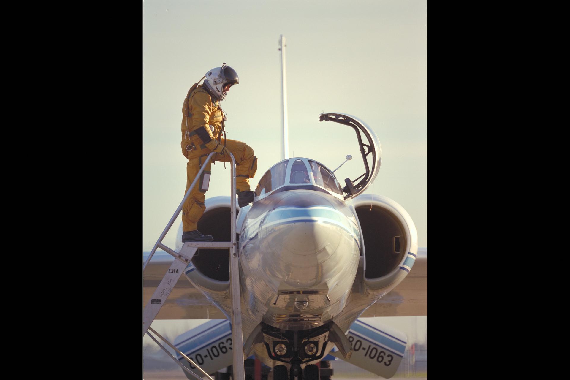 A pilot enters the cockpit of the ER-2 aircraft. He is outfitted with an oxygen suit with one foot stepping into the cockpit.