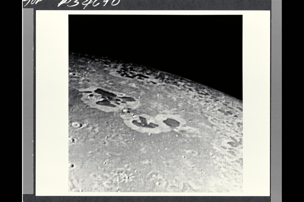 P-34690 Field of View : 1,000 km. or 600 miles This photograph of Neptune's moon Triton was acquired by Voyager 2. Triton's limb cuts obliquely accross the middle of the image. Three irregular dark areas, surrounded by brighter material, dominate the image. Low-lying material with intermediate albedo occupies the central area, anf fresh craters occur along the right margin. sub-parallel alignment of linear patches of dark material shown in the lower and left part of the image suggests that the patches are structurally controlled.