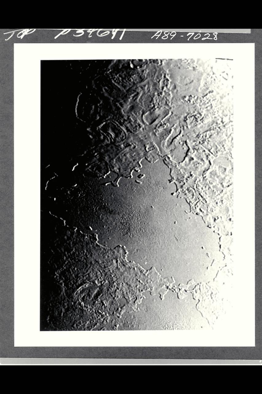 P-34668 This narrow angled image of Neptune's Triton, part of a sequence recieved from Voyager 2 shows a large flooded basin about 200 km. or 120 miles wide and 400 km. or 240 miles long. It appears to have been formed chiefly by two large, ancient craters. The walls of the basin have retreated, possibly as a consequence of sapping or collapse into the fluid that filled the main floor. The vent from which the flood evidently erupted lies near the right hand end of the basin. The surface of the eruptive material is rough near the vent and along the center of the basin, perhaps as a consequence of the presence of large rafts of ice. The absence of resolved impact craters on the floor of the basin indicatyes a young age for the flood.