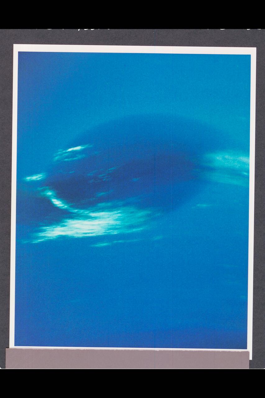 P-34668 Range :  2.8 million km. ( 1.7 million miles ) Smallest Resolvable Feature :  50 km or 31 miles This Voyager 2 image shows the last face on view of the Great Dark Spot that Voyager 2 will take with the narrow angle camera. It was made 45 hours before the closest approach to Neptune. the image shows feathery white clouds that overlie the boundary of the dark and light blue regions. the pinwheel ( spiral ) structure of both the dark boundary and the white cirrus suggest a storm system rotating counterclockwise. Periodic, small-scale patterns in the white clouds, possibly waves,  are short-lived and do not persist from one Neptunian rotation to the next. This color composite was made from the clear and green filter images.