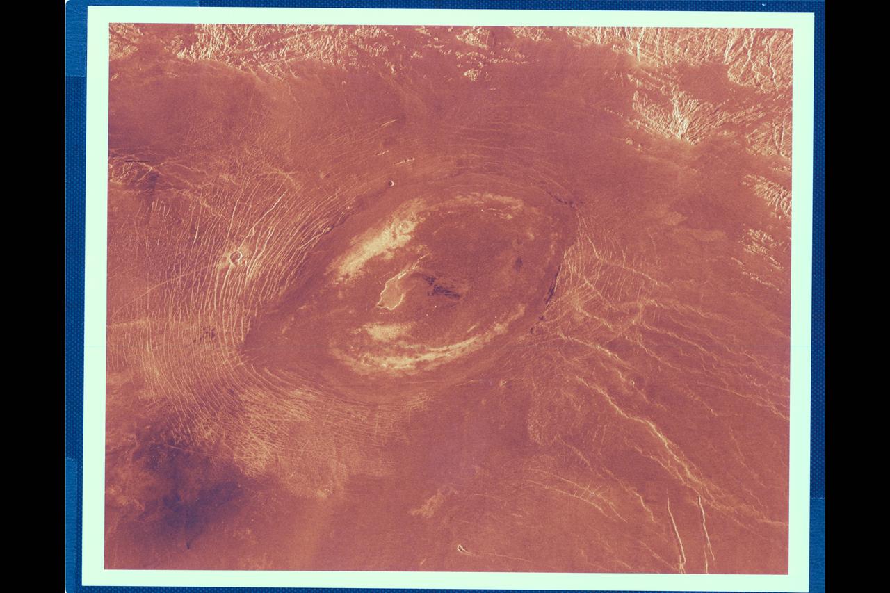 This Magellan image reveals Sacajawea Patera, a large, elongate caldera located in wester Ishtar Terra on the smooth plateau of Lakshmi Planum. The image is centered at 64.5 degrees north latitude adn 337 degrees east longitude. It is approx. 420 km (252 mi.) wide at the base Sacajawea is a depression approx. 1-2 km (0.6-1.2 mi.) deep and 120 by 215 km (74 by 133 mi.) in diameter; it is elongate in a sousthwest-northeast direction. The depression is interpreted to be graben adn fault scarps. These structures are space 0.5 to 4 km (0.3 to 2.5 mi.) apart, are 0.6 to 4 km (0.4 to 2.5 mi.) in width and up to 100 km (62 mi.) in length. Extending up to 140 km (87 mi.) in length from the southeast of the patera is a system of linear structures thought to represent a flanking rift zone along which the lateral injection and eruption of magma may have occurred. A shield edifice 12 km (7 mi.) in diameter with a prominent central pit lies along the trend of one of these features. The impact crater zlata, approx. 6 km (4 mi.) in diameter is located within the zone of graben to the northwest of the patera. Few flow features are observed in association with sacajawea, possibly due to age and state of degradation of the flows. Mottled bright deposits 4 to 20 km (2.5 to 12 mi.) in width are located near the periphery and in the center of the patera floor within local topographic lows. Diffuse patches of dark material approx. 40 km (25 mi.) in width are observed southwest of the patera, superimposed on portions of the surrounding graben. The formation of sacajawea is thought to be related to the drainage and collapse of a large magma chamber. Gravitational relaxation may have caused the resultant caldera to sag, producing the numerous complex, highly deformed tessera-like terrain are located north and east of the patera and are seen in the upper portion of the image. Color has been added to this image to simulate the appearance of the Venus surface.