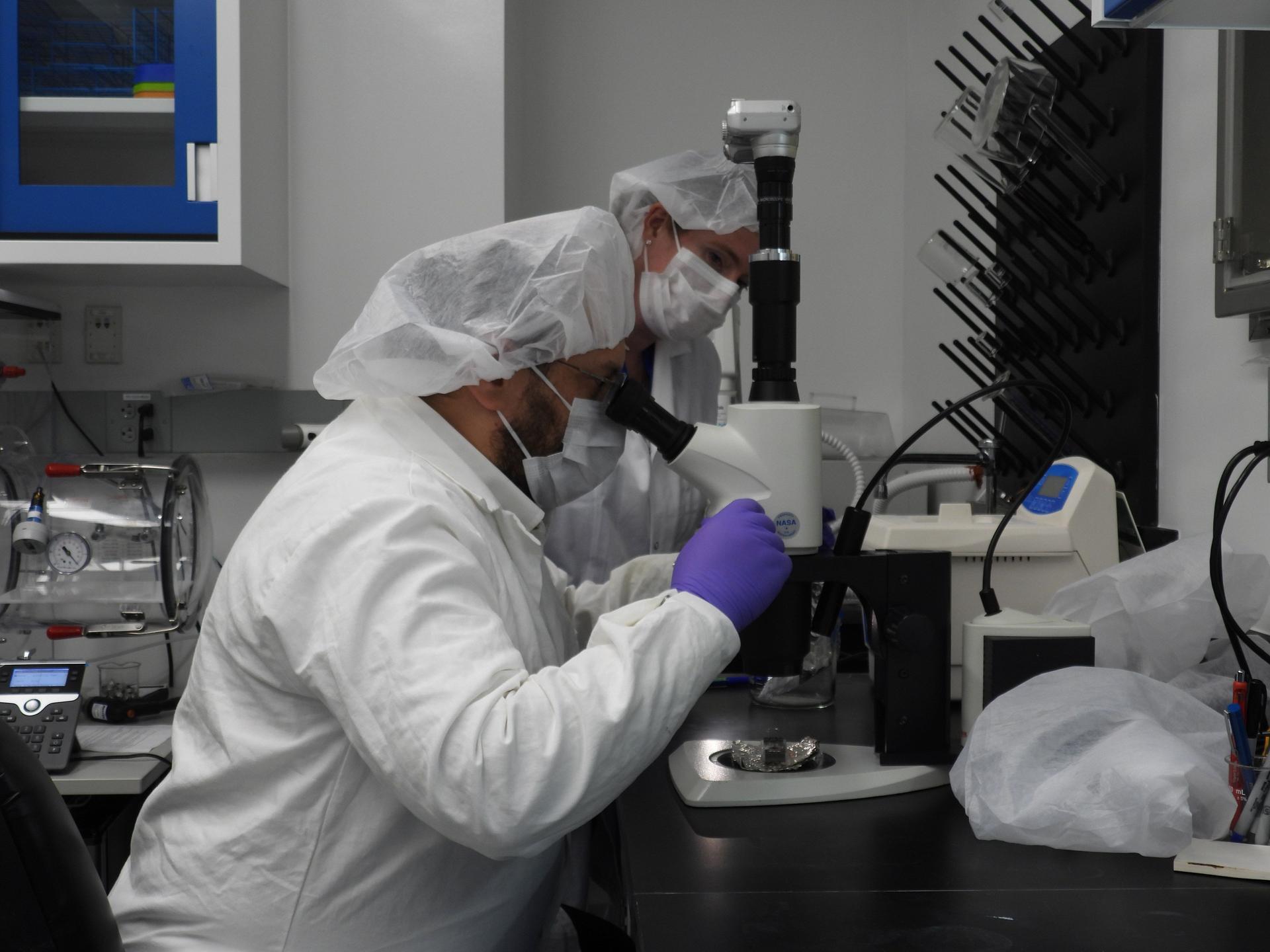 A man sits, looking into a microscope. He has on a white lab coat, purple gloves, and a white hair covering. Another person is wearing similar protective gear behind him.