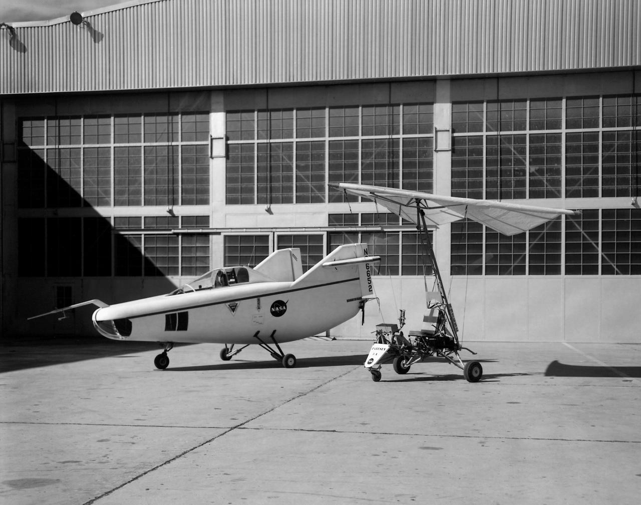 In this photo of the M2-F1 lifting body and the Paresev 1B on the ramp, the viewer sees two vehicles representing different approaches to building a research craft to simulate a spacecraft able to land on the ground instead of splashing down in the ocean as the Mercury capsules did. The M2-F1 was a lifting body, a shape able to re-enter from orbit and land. The Paresev (Paraglider Research Vehicle) used a Rogallo wing that could be (but never was) used to replace a conventional parachute for landing a capsule-type spacecraft, allowing it to make a controlled landing on the ground.
