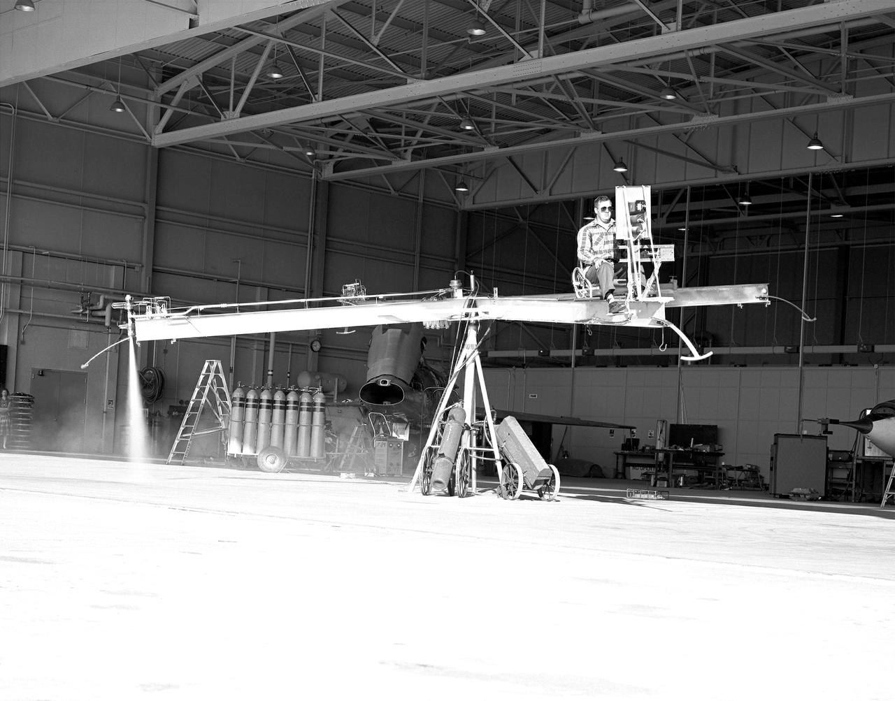 NACA High-Speed Flight Station test pilot Stan Butchart flying the Iron Cross, the mechanical reaction control simulator. High-pressure nitrogen gas expanded selectively, by the pilot, through the small reaction control thrusters maneuvered the Iron Cross through the three axes. The exhaust plume can be seen from the aft thruster. The tanks containing the gas can be seen on the cart at the base of the pivot point of the Iron Cross. NACA technicians built the iron-frame simulator, which matched the inertia ratios of the Bell X-1B airplane, installing six jet nozzles to control the movement about the three axes of pitch, roll, and yaw.