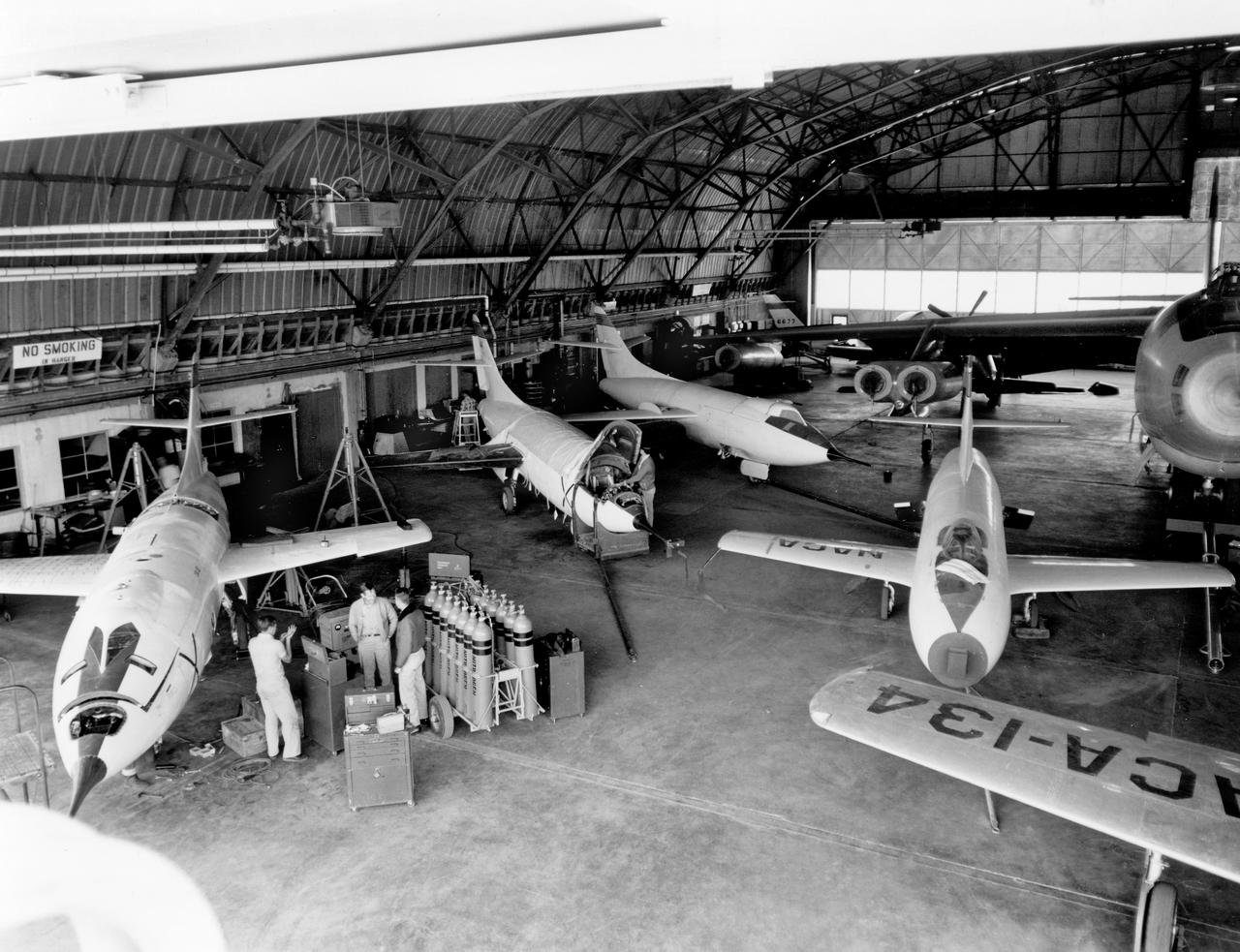 The aircraft in this 1953 photo of the National Advisory Committee for Aeronautics (NACA) hangar at South Base of Edwards Air Force Base showed the wide range of research activities being undertaken.  On the left side of the hangar are the three D-558-2 research aircraft. These were designed to test swept wings at supersonic speeds approaching Mach 2. The front D-558-2 is the third built (NACA 145/Navy 37975). It has been modified with a leading-edge chord extension. This was one of a number of wing modifications, using different configurations of slats and/or wing fences, to ease the airplane's tendency to pitch-up. NACA 145 had both a jet and a rocket engine. The middle aircraft is NACA 144 (Navy 37974), the second built. It was all-rocket powered, and Scott Crossfield made the first Mach 2 flight in this aircraft on November 20, 1953. The aircraft in the back is D-558-2 number 1. NACA 143 (Navy 37973) was also carried both a jet and a rocket engine in 1953. It had been used for the Douglas contractor flights, then was turned over to the NACA. The aircraft was not converted to all-rocket power until June 1954. It made only a single NACA flight before NACA's D-558-2 program ended in 1956.  Beside the three D-558-2s is the third D-558-1. Unlike the supersonic D-558-2s, it was designed for flight research at transonic speeds, up to Mach 1. The D-558-1 was jet-powered, and took off from the ground. The D-558-1's handling was poor as it approached Mach 1. Given the designation NACA 142 (Navy 37972), it made a total of 78 research flights, with the last in June 1953.  In the back of the hangar is the X-4 (Air Force 46-677). This was a Northrop-built research aircraft which tested a swept wing design without horizontal stabilizers. The aircraft proved unstable in flight at speeds above Mach 0.88. The aircraft showed combined pitching, rolling, and yawing motions, and the design was considered unsuitable. The aircraft, the second X-4 built, was then used as a pilot traine