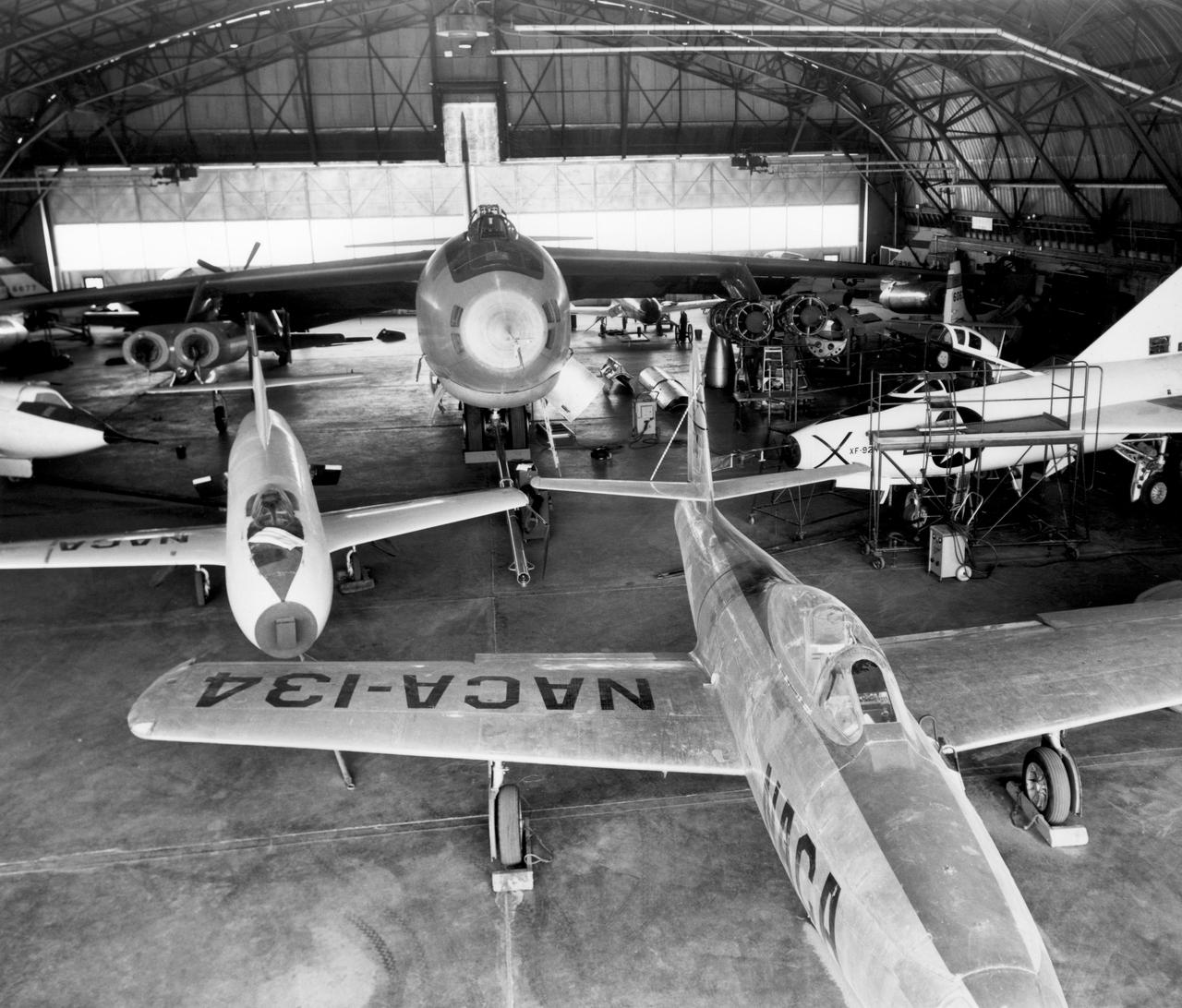 In the center foreground of this 1953 hangar photo is the YF-84A (NACA 134/Air Force 45-59490) used for vortex generator research. It arrived on November 28, 1949, and departed on April 21, 1954. Beside it is the third D-558-1 aircraft (NACA 142/Navy 37972). This aircraft was used for a total of 78 transonic research flights from April 1949 to June 1954. It replaced the second D-558-1, lost in the crash which killed Howard Lilly. Just visible on the left edge is the nose of the first D-558-2 (NACA 143/Navy 37973). Douglas turned the aircraft over to NACA on August 31, 1951, after the contractor had completed its initial test flights. NACA only made a single flight with the aircraft, on September 17, 1956, before the program was cancelled. In the center of the photo is the B-47A (NACA 150/Air Force 49-1900). The B-47 jet bomber, with its thin, swept-back wings, and six podded engines, represented the state of the art in aircraft design in the early 1950s. The aircraft undertook a number of research activities between May 1953 and its 78th and final research flight on November 22, 1957. The tests showed that the aircraft had a buffeting problem at speeds above Mach 0.8. Among the pilots who flew the B-47 were later X-15 pilots Joe Walker, A. Scott Crossfield, John B. McKay, and Neil A. Armstrong.  On the right side of the B-47 is NACA's X-1 (Air Force 46-063). The second XS-1 aircraft built, it was fitted with a thicker wing than that on the first aircraft, which had exceeded Mach 1 on October 14, 1947. Flight research by NACA pilots indicated that this thicker wing produced 30 percent more drag at transonic speeds compared to the thinner wing on the first X-1. After a final flight on October 23, 1951, the aircraft was grounded due to the possibility of fatigue failure of the nitrogen spheres used to pressurize the fuel tanks. At the time of this photo, in 1953, the aircraft was in storage. In 1955, the aircraft was extensively modified, becoming the X-1E.  In front o