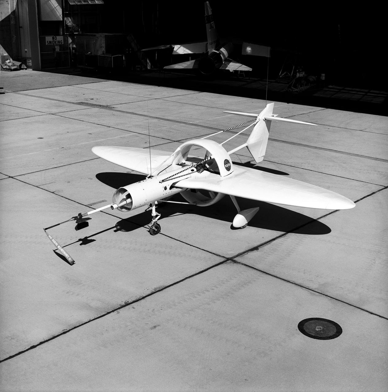 This 1976 photograph of the Oblique Wing Research Aircraft was taken in front of the NASA Flight Research Center hangar, located at Edwards Air Force Base, California. In the photograph the noseboom, pitot-static probe, and angles-of-attack and sideslip flow vanes(covered-up) are attached to the front of the vehicle. The clear nose dome for the television camera, and the shrouded propellor for the 90 horsepower engine are clearly seen.