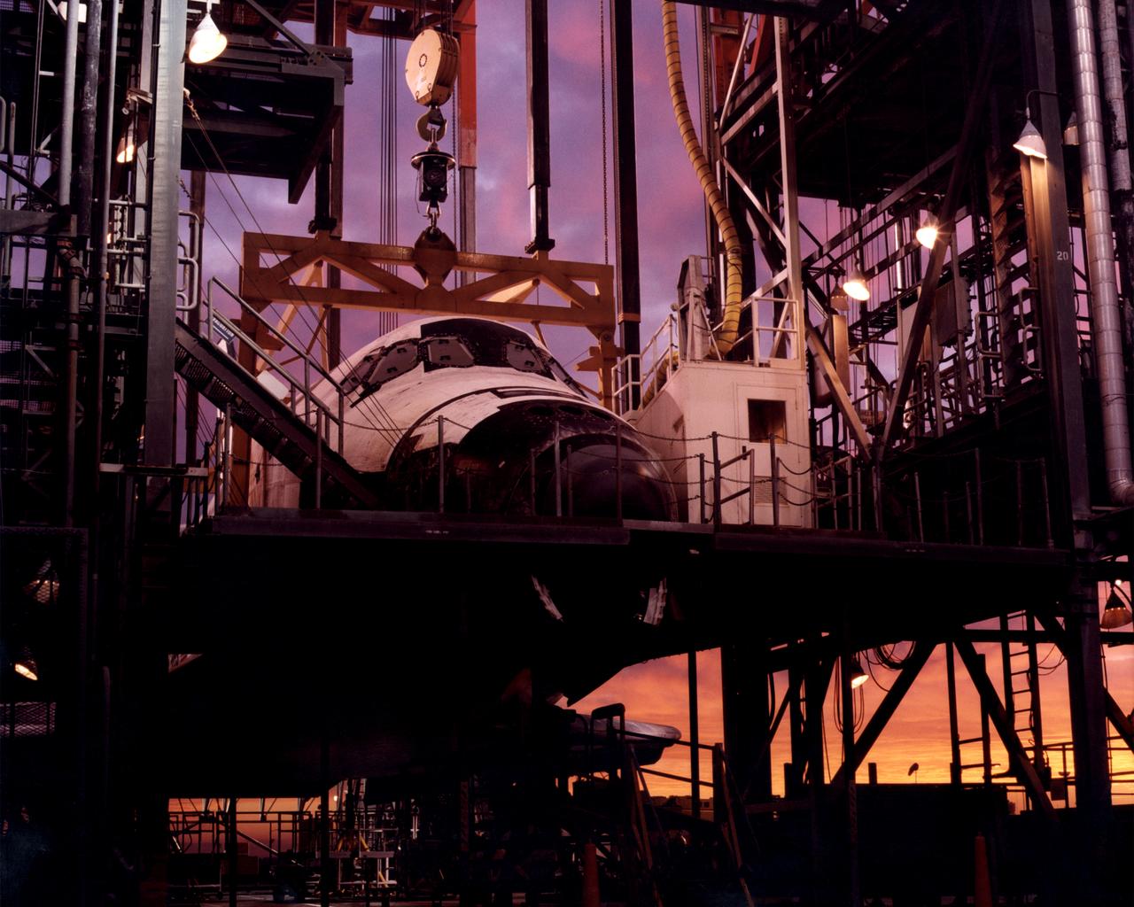 The early-morning Sun bathes the Space Shuttle Discovery in hues of purple, pink and gold as it is encased in the Mate-Demate Device (MDD) at NASA’s Dryden Flight Research Center at Edwards, California. The gantry-like MDD structure is used to prepare the shuttle for its ferry flight back to the Kennedy space Center in Florida, including mounting the shuttle atop NASA’s modified Boeing 747 Shuttle Carrier Aircraft.