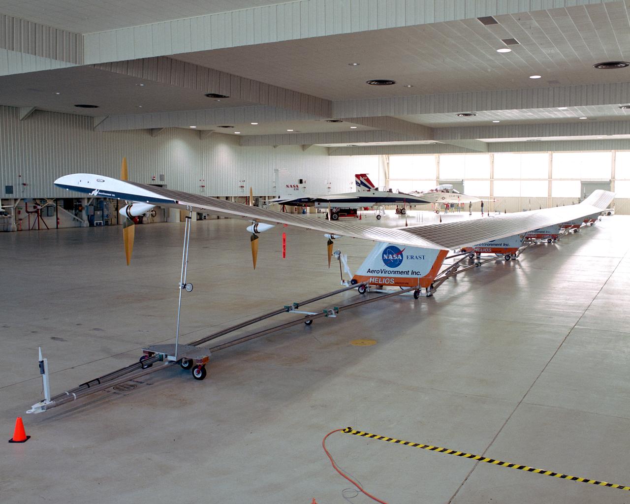 The solar-powered Helios Prototype flying wing frames two modified F-15 research aircraft in a hangar at NASA's Dryden Flight Research Center, Edwards, California. The elongated 247-foot span lightweight aircraft, resting on its ground maneuvering dolly, stretched almost the full length of the 300-foot long hangar while on display during a visit of NASA Administrator Sean O'Keefe and other NASA officials on Jan. 31, 2002. The unique solar-electric flying wing reached an altitude of 96,863 feet during an almost 17-hour flight near Hawaii on Aug. 13, 2001, a world record for sustained horizontal flight by a non-rocket powered aircraft. Developed by AeroVironment, Inc., under NASA's Environmental Research Aircraft and Sensor Technology (ERAST) project, the Helios Prototype is the forerunner of a planned fleet of slow-flying, long duration, high-altitude uninhabited aerial vehicles (UAV) which can serve as "atmospheric satellites," performing Earth science missions or functioning as telecommunications relay platforms in the stratosphere.