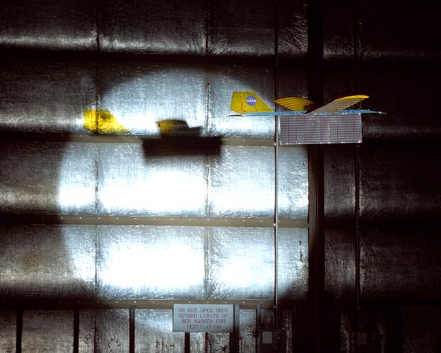 An experimental radio-controlled model aircraft casts a unique shadow as it flies inside a Dryden hangar using only a spotlight as an energy source.