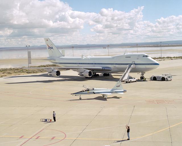 NASA image: White clouds and a flooded lakebed form a backdrop as a T-38 support aircraft taxies across the ramp in front of NASA's Boeing 747 Shuttle Carrier Aircraft