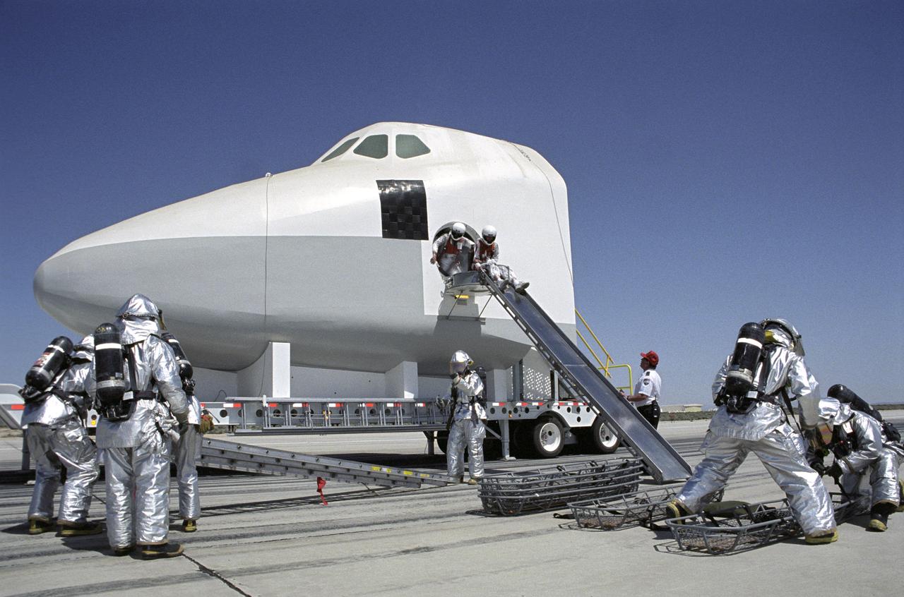 While fire-rescue personnel prepare evacuation litters, two stand-in "astronauts" prepare to use an exit slide from a Shuttle mockup during a rescue training exercise.