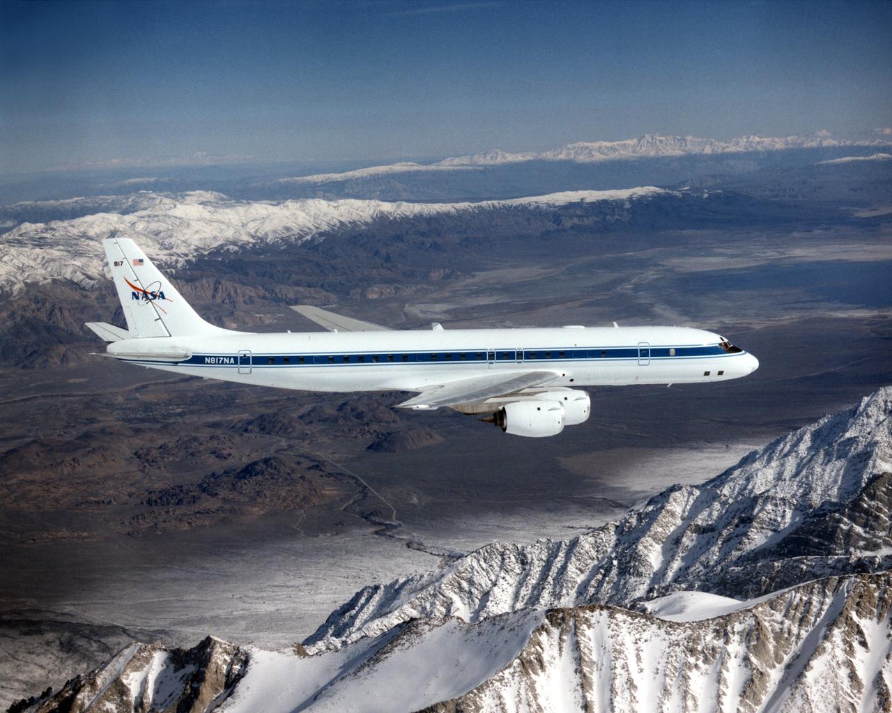 The DC-8 in flight near Lone Pine, Calif. In the foreground are the Sierra Nevada Mountains, covered with winter snow. In the distance are the White Mountains. The DC-8's fuselage is painted white with a dark blue stripe down the side. The wings are silver, while the engine pods are white. In this view of the airplane's right-hand side, only a few of its antennas are visible. The experimental payload can be as great as 30,000 pounds of equipment for gathering data of various sorts.