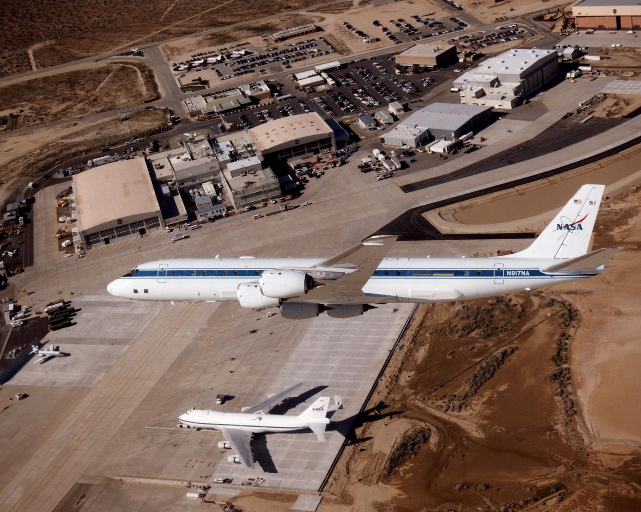 The DC-8 aircraft is seen making a banking turn high above the NASA Dryden ramp. This view of the DC-8's left side reveals some of the modifications necessary for particular on-board experiments. To the right of the DC-8 is the edge of Rogers Dry Lake. Above the aircraft's forward fuselage is the Dryden Flight Research Center headquarters building, while other NASA facilities extend down the flightline to the right. Below the DC-8 is the Shuttle Carrier Aircraft (SCA), on which are visible attachment points for the Shuttle Orbiter.