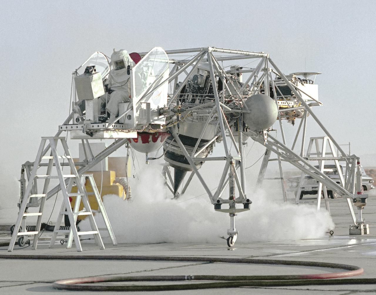This 1964 NASA Flight Reserch Center photograph shows a ground engine test underway on the Lunar Landing Research Vehicle (LLRV) number 1. When Apollo planning was underway in 1960, NASA was looking for a simulator to profile the descent to the Moon's surface. Three concepts surfaced: an electronic simulator, a tethered device, and the ambitious Dryden contribution, a free-flying vehicle. All three became serious projects, but eventually the NASA Flight Research Center's (FRC) Landing Research Vehicle (LLRV) became the most significant one. Hubert M. Drake is credited with originating the idea, while Donald Bellman and Gene Matranga were senior engineers on the project, with Bellman, the project manager. Simultaneously, and independently, Bell Aerosystems Company, Buffalo, N.Y., a company with experience in vertical takeoff and landing (VTOL) aircraft, had conceived a similar free-flying simulator and proposed their concept to NASA headquarters. NASA Headquarters put FRC and Bell together to collaborate. The challenge was; to allow a pilot to make a vertical landing on Earth in a simulated Moon environment, one sixth of the Earth's gravity and with totally transparent aerodynamic forces in a "free flight" vehicle with no tether forces acting on it.  Built of tubular aluminum like a giant four-legged bedstead, the vehicle was to simulate a lunar landing profile from around 1500 feet to the Moon's surface. To do this, the LLRV had a General Electric CF-700-2V turbofan engine mounted vertically in gimbals, with 4200 pounds of thrust. The engine, using JP-4 fuel, got the vehicle up to the test altitude and was then throttled back to support five-sixths of the vehicle's weight, simulating the reduced gravity of the Moon. Two hydrogen-peroxide lift rockets with thrust that could be varied from 100 to 500 pounds handled the LLRV's rate of descent and horizontal translations. Sixteen smaller hydrogen-peroxide rockets, mounted in pairs, gave the pilot control in pitch, yaw, 
