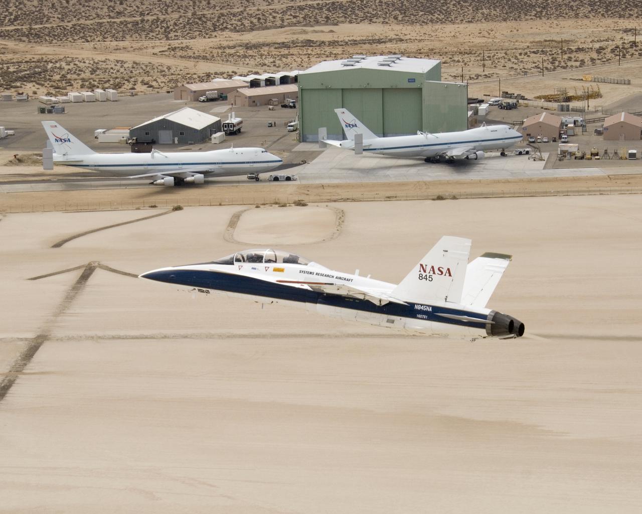 NASA's two Boeing 747 Shuttle Carrier Aircraft form the backdrop as pilot Dick Ewers banks NASA F/A-18 #845 low over Rogers Dry Lake to end a research flight.