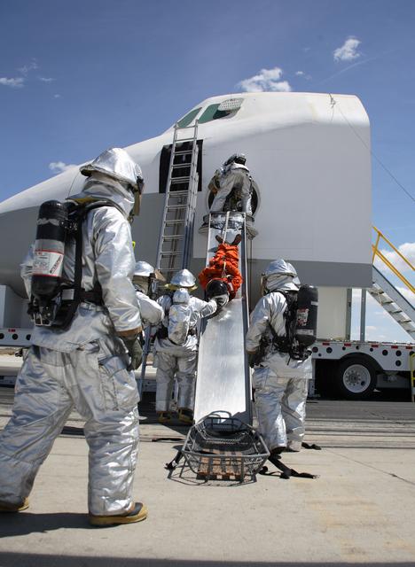 Clad in thermal protection suits, fire/rescue crew aid a volunteer "Injured astronaut" to a head-first ride down the exit slide from the shuttle cabin mockup