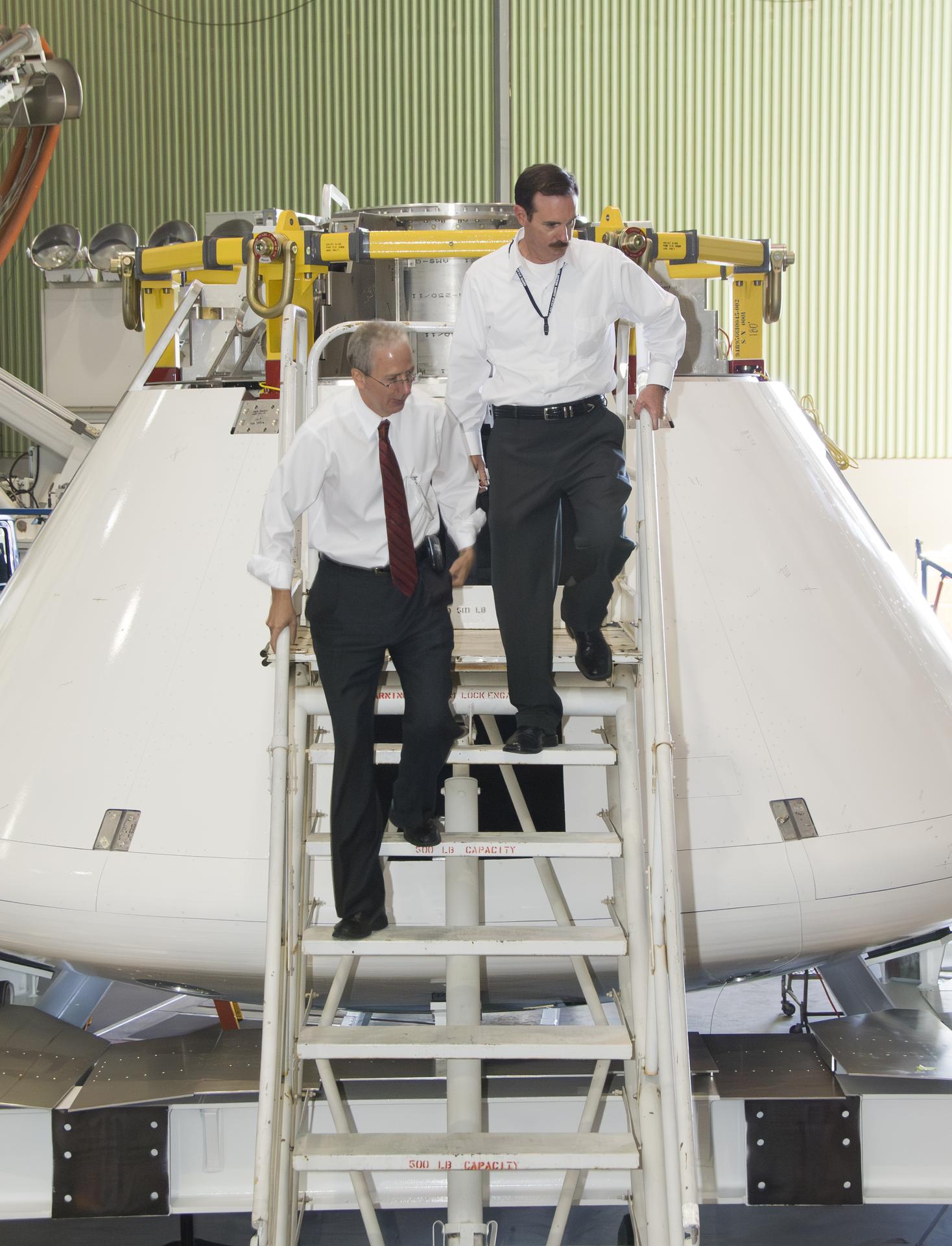 Two men walk down a set of stairs from a mock-up of a space capsule.