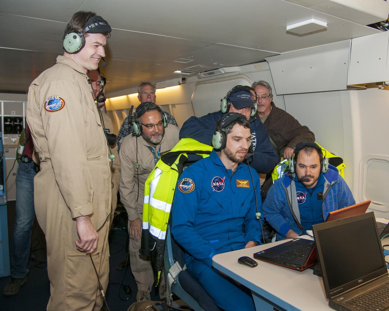Scientists onboard SOFIA watch data come in from Pluto’s atmosphere as SOFIA flies into the shadow the dwarf planet cast on Earth’s surface during an eclipse-like event called an occultation. (Left to right standing): Holger Jakob, Thomas Roellig, (unknown who is hunched over), Jürgen Wolf, Nicholas Veronico (Left to right seated): Enrico Pfüller, Manuel Wiedemann