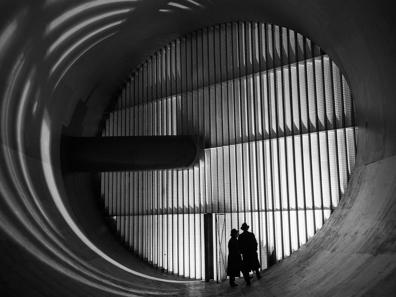 Men stand in front of turning vanes inside the Altitude Wind Tunnel (AWT) at the National Advisory Committee for Aeronautics (NACA) Aircraft Engine Research Laboratory. The AWT was the only wind tunnel capable of testing full-size aircraft engines in simulated altitude conditions. A large wooden drive fan, located on the other side of these vanes, created wind speeds up to 500 miles per hour. The drive shaft connected the fan to the induction motor located in an adjacent building.        Turning vanes were located in each corner of the rectangular tunnel to straighten the airflow and direct it around the corners. This set of vanes was located in the 31-foot-diameter southeast corner of the tunnel. These elliptical panels consisted of 36 to 42 vertical vanes that were supported by three horizontal supports. The individual vanes were 2.5 feet long and half-moon shaped. The panel of vanes was affixed to the curved corner rings of the tunnel. Each set of turning vanes had a moveable vane in the middle of the lower level for personnel access.    Each set of vanes took weeks to assemble before they were installed during the summer of 1943. This publicity photograph was taken just weeks after the tunnel became operational in February 1944.