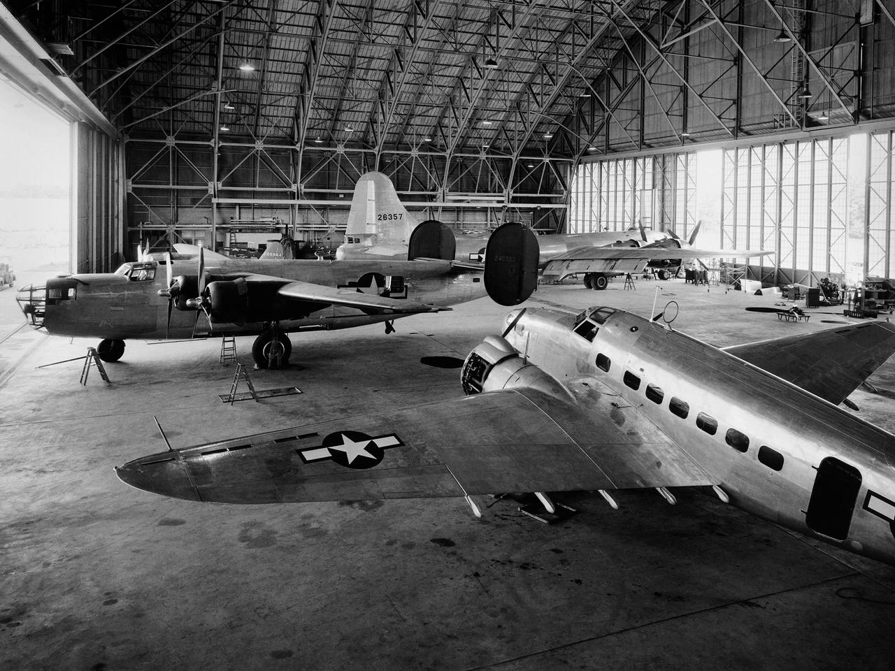 A Consolidated B–24D Liberator (left), Boeing B–29 Superfortress (background), and Lockheed RA–29 Hudson (foreground) parked inside the Flight Research Building at the National Advisory Committee for Aeronautics (NACA) Aircraft Engine Research Laboratory in Cleveland, Ohio. A P–47G Thunderbolt and P–63A King Cobra are visible in the background. The laboratory utilized 15 different aircraft during the final 2.5 years of World War II. This starkly contrasts with the limited-quantity, but long-duration aircraft of the NASA’s modern fleet.       The Flight Research Building is a 272- by 150-foot hangar with an internal height ranging from 40 feet at the sides to 90 feet at its apex. The steel support trusses were pin-connected at the top with tension members extending along the corrugated transite walls down to the floor. The 37.5-foot-tall and 250-foot-long doors on either side can be opened in sections. The hangar included a shop area and stock room along the far wall, and a single-story office wing with nine offices, behind the camera. The offices were later expanded.    The hangar has been in continual use since its completion in December 1942. Nearly 70 different aircraft have been sheltered here over the years. Temporary offices were twice constructed over half of the floor area when office space was at a premium.
