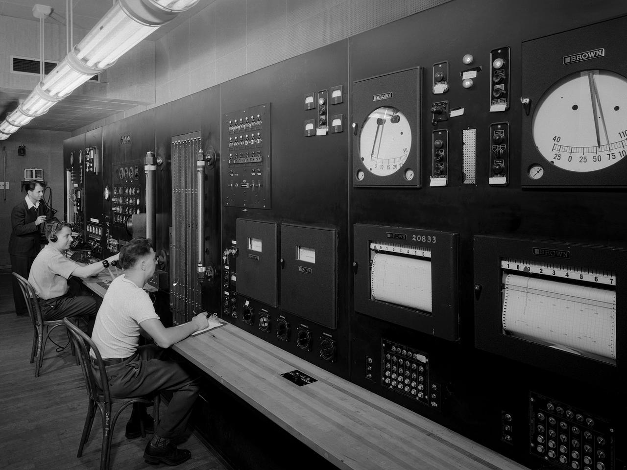 Operators in the control room for the Altitude Wind Tunnel at the National Advisory Committee for Aeronautics (NACA) Aircraft Engine Research Laboratory remotely operate a Wright R–3350 engine in the tunnel’s test section. Four of the engines were used to power the B–29 Superfortress, a critical weapon in the Pacific theater during World War II. The wind tunnel, which had been in operation for approximately six months, was the nation’s only wind tunnel capable of testing full-scale engines in simulated altitude conditions.        The soundproof control room was used to operate the wind tunnel and control the engine being run in the test section. The operators worked with assistants in the adjacent Exhauster Building and Refrigeration Building to manage the large altitude simulation systems. The operator at the center console controlled the tunnel’s drive fan and operated the engine in the test section. Two sets of pneumatic levers near his right forearm controlled engine fuel flow, speed, and cooling. Panels on the opposite wall, out of view to the left, were used to manage the combustion air, refrigeration, and exhauster systems.    The control panel also displayed the master air speed, altitude, and temperature gauges, as well as a plethora of pressure, temperature, and airflow readings from different locations on the engine. The operator to the right monitored the manometer tubes to determine the pressure levels. Despite just being a few feet away from the roaring engine, the control room remained quiet during the tests.
