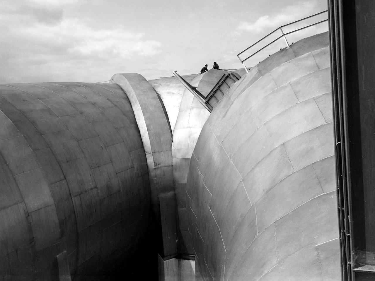 Two men on top of the Altitude Wind Tunnel (AWT) at the National Advisory Committee for Aeronautics (NACA) Aircraft Engine Research Laboratory. The tunnel was a massive rectangular structure, which for years provided one of the highest vantage points on the laboratory. The tunnel was 263 feet long on the north and south legs and 121 feet long on the east and west sides. The larger west end of the tunnel, seen here, was 51 feet in diameter. The east side of the tunnel was 31 feet in diameter at the southeast corner and 27 feet in diameter at the northeast. The throat section, which connected the northwest corner to the test section, narrowed sharply from 51 to 20 feet in diameter.       The AWT’s altitude simulation required temperature and pressure fluctuations that made the design of the shell more difficult than other tunnels. The simultaneous decrease in both pressure and temperature inside the facility produced uneven stress loads, particularly on the support rings. The steel used in the primary tunnel structure was one inch thick to ensure that the shell did not collapse as the internal air pressure was dropped to simulate high altitudes. It was a massive amount of steel considering the World War II shortages.    The shell was covered with several inches of fiberglass insulation to retain the refrigerated air and a thinner outer steel layer to protect the insulation against the weather. A unique system of rollers was used between the shell and its support piers. These rollers allowed for movement as the shell expanded or contracted during the altitude simulations. Certain sections would move as much as five inches during operation.