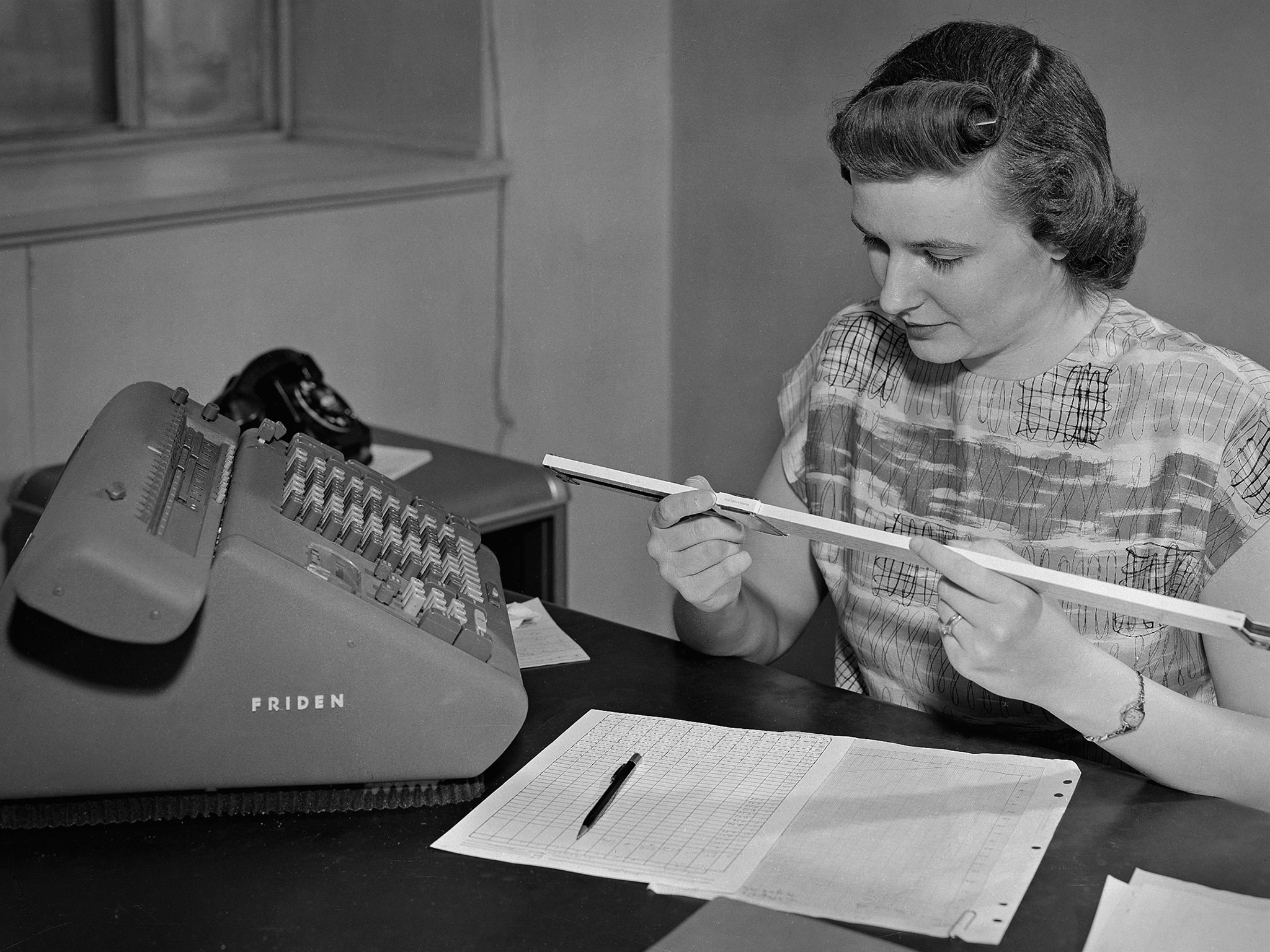 A woman uses a slide rule and Friden machine at a desk
