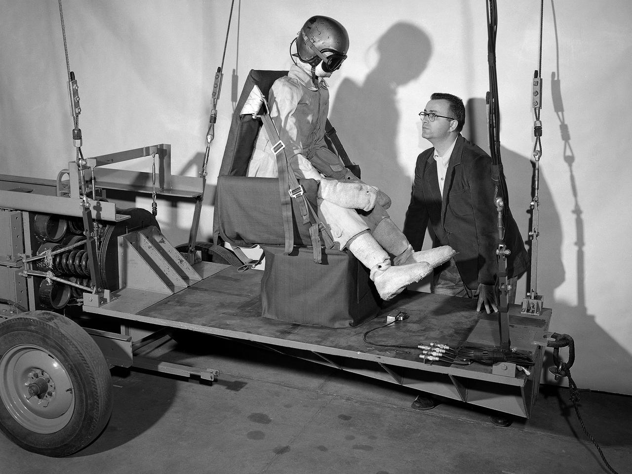 A researcher at the National Advisory Committee for Aeronautics (NACA) Lewis Flight Propulsion Laboratory prepares for a test of an NACA-designed aircraft seat. The laboratory had undertaken a multi-year investigation into the causes and prevention of fires on low altitude aircraft crashes. The program was expanded in the mid-1950s to include the study of impact on passengers, types of seat restraints, and seat design.      The crash impact portion of the program began by purposely wrecking surplus Fairchild C-82 Packet and Piper Cub aircraft into barricades at the end of a test runway at the Ravenna Arsenal, located approximately 40 miles south of the Lewis lab in Cleveland. Instrumented dummies and cameras were installed in the pilot and passenger areas. After determining the different loads and their effects on the passengers, the NACA researchers began designing new types of seats and restraints.     The result was an elastic seat that flexed upon impact, absorbing 75 percent of the loads before it slowly recoiled. This photograph shows the seats mounted on a pendulum with a large spring behind the platform to provide the jolt that mimicked the forces of a crash. The seat was constructed without any potentially damaging metal parts and included rubber-like material, an inflated back and arms, and a seat cushion. After the pendulum tests, the researchers compared the flexible seats to the rigid seats during a crash of a transport aircraft. They found the passengers in the rigid seats received 66 percent higher g-forces than the NACA-designed seats.