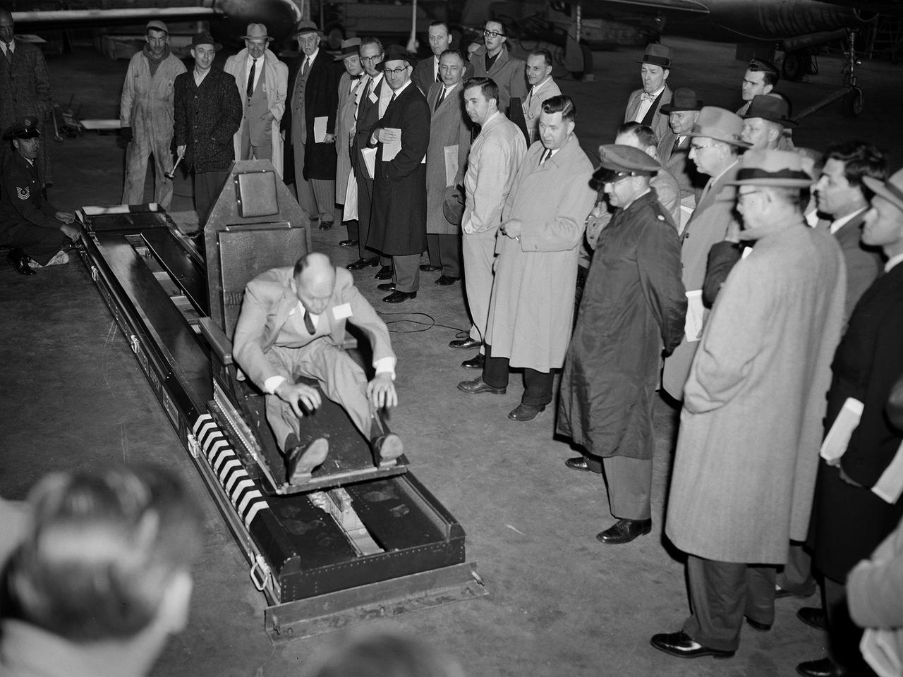 Howard Hasbrook volunteers for a demonstration of a scaled-down version of Lieutenant Colonel John Stapp’s rocket sled set up in the hangar at the National Advisory Committee for Aeronautics (NACA) Lewis Flight Propulsion Laboratory. In 1945 Stapp, an Air Force medical doctor, volunteered to participate in a deceleration program to study the human body’s tolerance to aircraft crash forces. A 1500-pound sled powered by rockets was installed in 1947 on a section of railroad track in the California desert. Stapp participated in 29 experiments over the next seven years and broke land and deceleration records. These tests studied the effects of acceleration, G-force, deceleration, and wind blast on humans. Stapp suffered broken bones and retinal hemorrhages, but suffered no permanent damage.     NACA Lewis was conducting a series of crash impact studies in the mid-1950s using dummies in actual aircraft. Irving Pinkel, the director of the program, and Stapp became friends through their mutual interest in this field. In April 1956 Stapp visited the Cleveland lab to give a talk to the local section of the American Rocket Society that discussed issues relating to the escape of pilots from the cockpit of supersonic jet aircraft. That same week, NACA Lewis’ Pinkel, Gerard Pesman, Merritt Preston, and Dugald Black received the annual Laura Taber Barbour Air Safety Award for their work on the Crash Fire Program. Black and Preston are visible in the crowd in this photograph.
