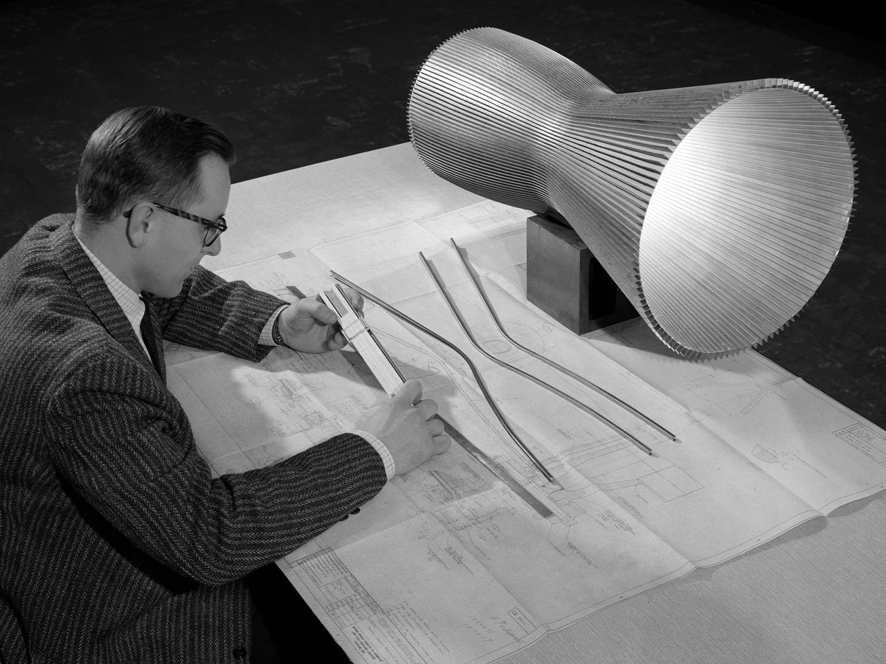 An engineer at the National Aeronautics and Space Administration (NASA) Lewis Research Center examines a drawing showing the assembly and details of a 20,000-pound thrust regeneratively cooled rocket engine. The engine was being designed for testing in Lewis’ new Rocket Engine Test Facility, which began operating in the fall of 1957. The facility was the largest high-energy test facility in the country that was capable of handling liquid hydrogen and other liquid chemical fuels. The facility’s use of subscale engines up to 20,000 pounds of thrust permitted a cost-effective method of testing engines under various conditions.      The Rocket Engine Test Facility was critical to the development of the technology that led to the use of hydrogen as a rocket fuel and the development of lightweight, regeneratively-cooled, hydrogen-fueled rocket engines. Regeneratively-cooled engines use the cryogenic liquid hydrogen as both the propellant and the coolant to prevent the engine from burning up. The fuel was fed through rows of narrow tubes that surrounded the combustion chamber and nozzle before being ignited inside the combustion chamber. The tubes are visible in the liner sitting on the desk.    At the time, Pratt and Whitney was designing a 20,000-pound thrust liquid-hydrogen rocket engine, the RL-10. Two RL-10s would be used to power the Centaur second-stage rocket in the 1960s. The successful development of the Centaur rocket and the upper stages of the Saturn V were largely credited to the work carried out Lewis.