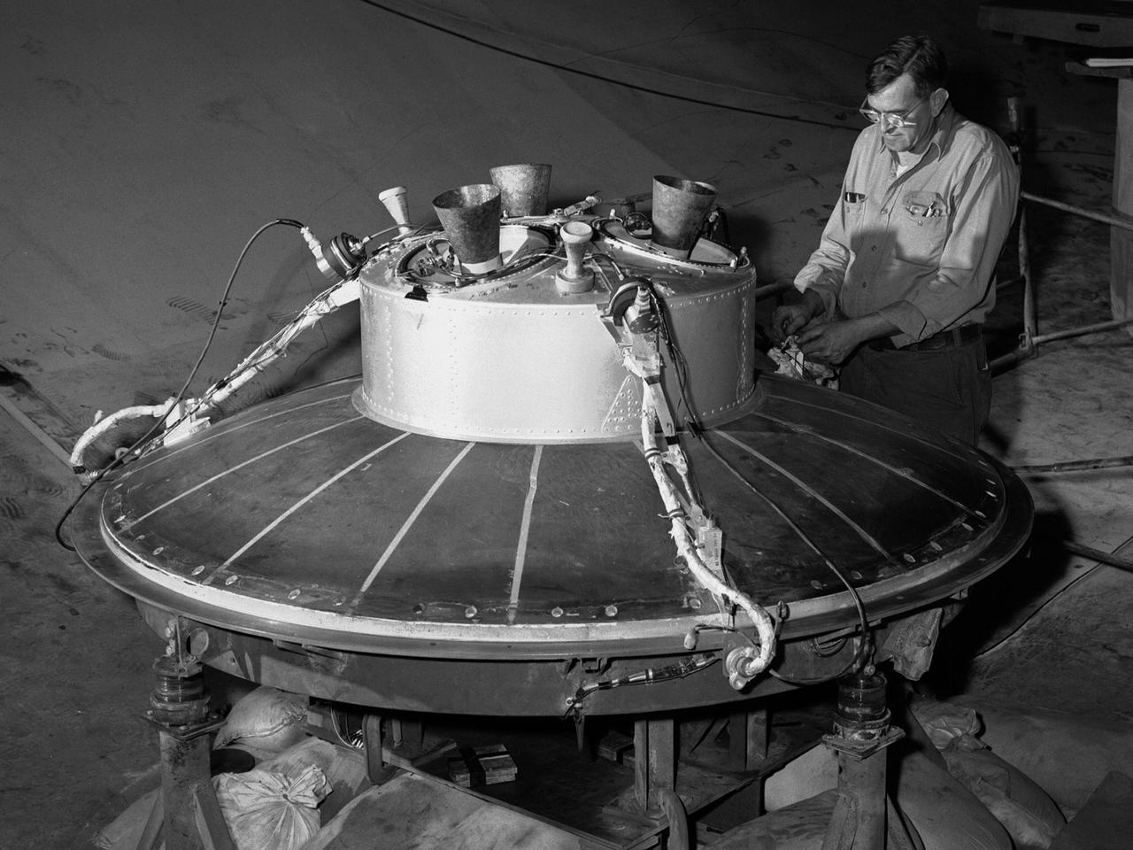 A mechanic at the National Aeronautics and Space Administration (NASA) Lewis Research Center prepares the inverted base of a Mercury capsule for a test of its posigrade retrorockets inside the Altitude Wind Tunnel. In October 1959 NASA’s Space Task Group allocated several Project Mercury assignments to Lewis. The Altitude Wind Tunnel was modified to test the Atlas separation system, study the escape tower rocket plume, train astronauts to bring a spinning capsule under control, and calibrate the capsule’s retrorockets. The turning vanes, makeup air pipes, and cooling coils were removed from the wide western end of the tunnel to create a 51-foot diameter test chamber.    The Mercury capsule had a six-rocket retro-package affixed to the bottom of the capsule. Three of these were posigrade rockets used to separate the capsule from the booster and three were retrograde rockets used to slow the capsule for reentry into the earth’s atmosphere. Performance of the retrorockets was vital since there was no backup system.     Qualification tests of the retrorockets began in April 1960 on a retrograde thrust stand inside the southwest corner of the Altitude Wind Tunnel. These studies showed that a previous issue concerning the delayed ignition of the propellant had been resolved. Follow-up test runs verified reliability of the igniter’s attachment to the propellant. In addition, the capsule’s retrorockets were calibrated so they would not alter the capsule’s attitude when fired.