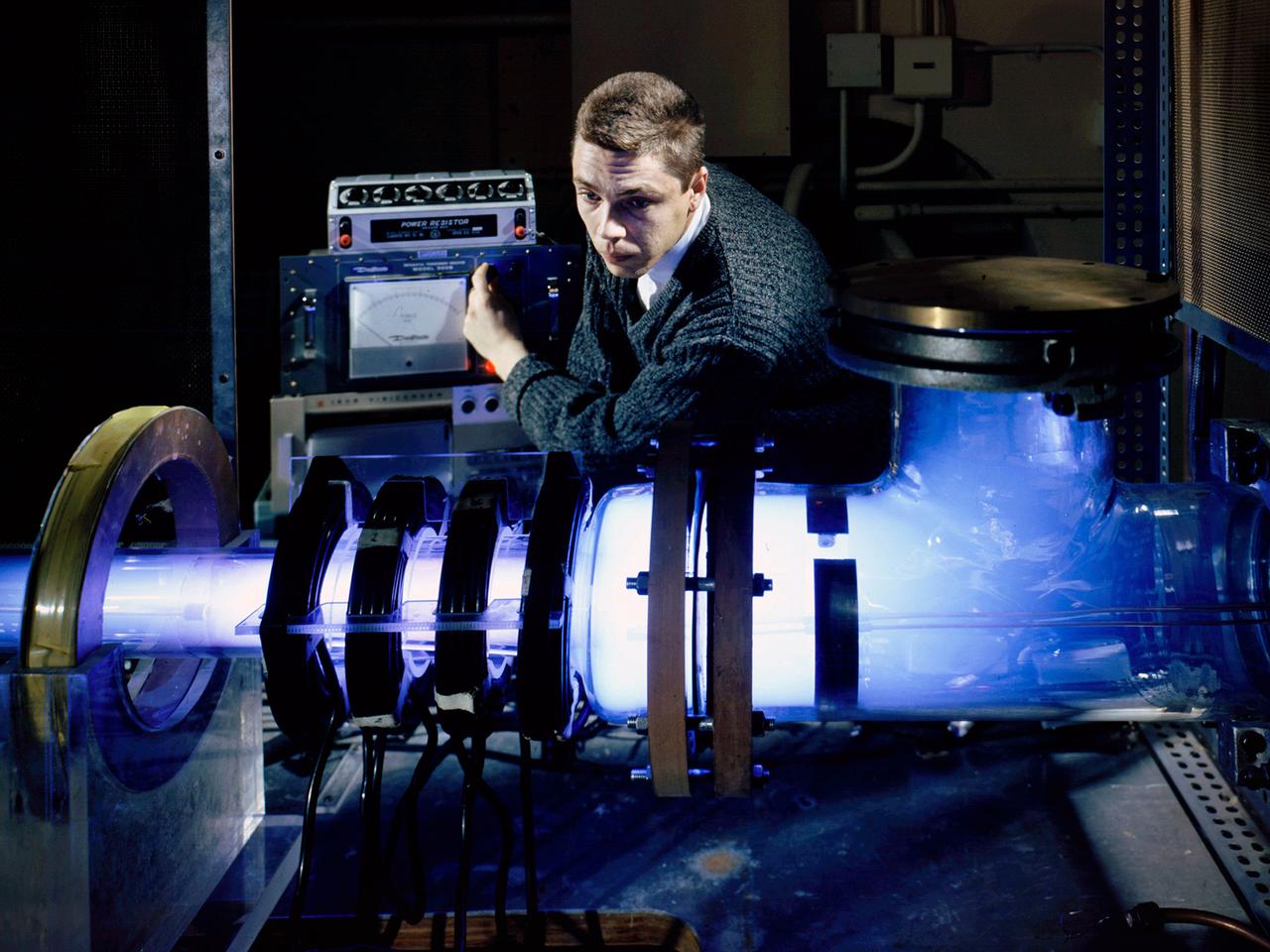 Raymond Palmer, of the Electromagnetic Propulsion Division’s Plasma Flow Section, adjusts the traveling magnetic wave plasma engine being operated in the Electric Power Conversion at the National Aeronautics and Space Administration (NASA) Lewis Research Center. During the 1960s Lewis researchers were exploring several different methods of creating electric propulsion systems, including the traveling magnetic wave plasma engine. The device operated similarly to alternating-current motors, except that a gas, not a solid, was used to conduct the electricity. A magnetic wave induced a current as it passed through the plasma. The current and magnetic field pushed the plasma in one direction.    Palmer and colleague Robert Jones explored a variety of engine configurations in the Electric Propulsion Research Building. The engine is seen here mounted externally on the facility’s 5-foot diameter and 16-foot long vacuum tank. The four magnetic coils are seen on the left end of the engine. The researchers conducted two-minute test runs with varying configurations and used of both argon and xenon as the propellant.   The Electric Propulsion Research Building was built in 1942 as the Engine Propeller Research Building, often called the Prop House. It contained four test cells to study large reciprocating engines with their propellers. After World War II, the facility was modified to study turbojet engines. By the 1960s, the facility was modified again for electric propulsion research and given its current name.