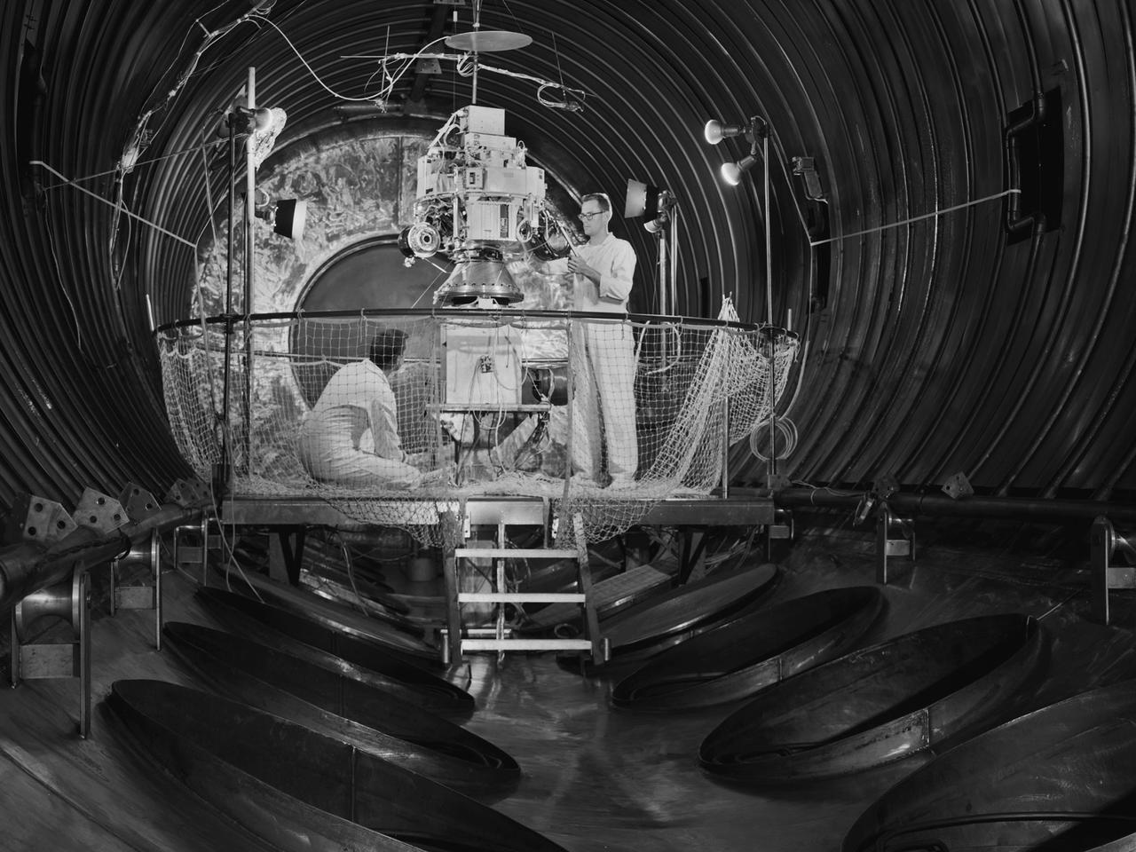 Technicians prepare the Space Electric Research Test (SERT-I) payload for a test in Tank Number 5 of the Electric Propulsion Laboratory at the National Aeronautics and Space Administration (NASA) Lewis Research Center. Lewis researchers had been studying different methods of electric rocket propulsion since the mid-1950s. Harold Kaufman created the first successful engine, the electron bombardment ion engine, in the early 1960s. These electric engines created and accelerated small particles of propellant material to high exhaust velocities. Electric engines have a very small amount of thrust, but once lofted into orbit by workhorse chemical rockets, they are capable of small, continuous thrust for periods up to several years.    The electron bombardment thruster operated at a 90-percent efficiency during testing in the Electric Propulsion Laboratory. The package was rapidly rotated in a vacuum to simulate its behavior in space. The SERT-I mission, launched from Wallops Island, Virginia, was the first flight test of Kaufman’s ion engine. SERT-I had one cesium engine and one mercury engine. The suborbital flight was only 50 minutes in duration but proved that the ion engine could operate in space.      The Electric Propulsion Laboratory included two large space simulation chambers, one of which is seen here. Each uses twenty 2.6-foot diameter diffusion pumps, blowers, and roughing pumps to remove the air inside the tank to create the thin atmosphere. A helium refrigeration system simulates the cold temperatures of space.