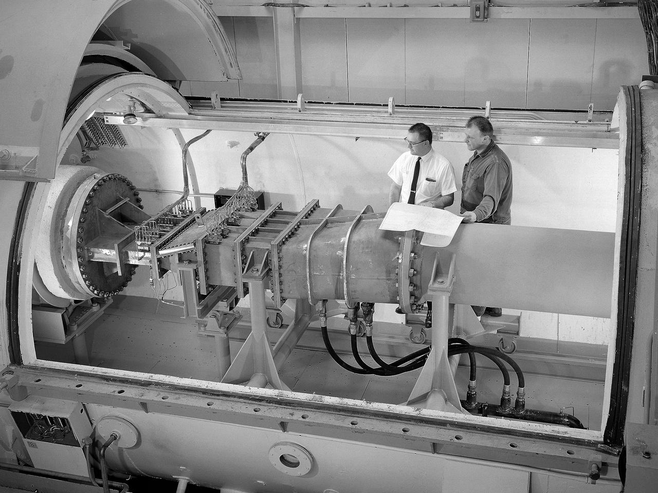 Engineer Frank Kutina and a National Aeronautics and Space Administration (NASA) mechanic examine the setup of an advanced combustor rig inside one of the test cells at the Lewis Research Center’s Four Burner Area in the Engine Research Building. Kutina, of the Research Operations Branch, served as go-between for the researchers and the mechanics. He helped develop the test configurations and get the hardware installed. At the time of this photograph, Lewis Center Director Abe Silverstein had just established the Airbreathing Engine Division to address the new propulsion of the 1960s. After nearly a decade of focusing almost exclusively on space, NASA Lewis began tackling issues relating to the new turbofan engine, noise reduction, energy efficiency, supersonic transport, and the never-ending quest for higher performance levels with smaller and more lightweight engines. The Airbreathing Engine Division’s Combustion Branch was dedicated to the study and mitigation of the high temperatures and pressures found in advanced combustor designs. These high temperatures and pressures could destroy engine components. The Lewis investigation included film cooling, diffuser flow, and jet mixing. Components were tested in smaller test cells, but a full-scale augmenting burner rig, seen here, was tested extensively in the Four Burner Area test cell.