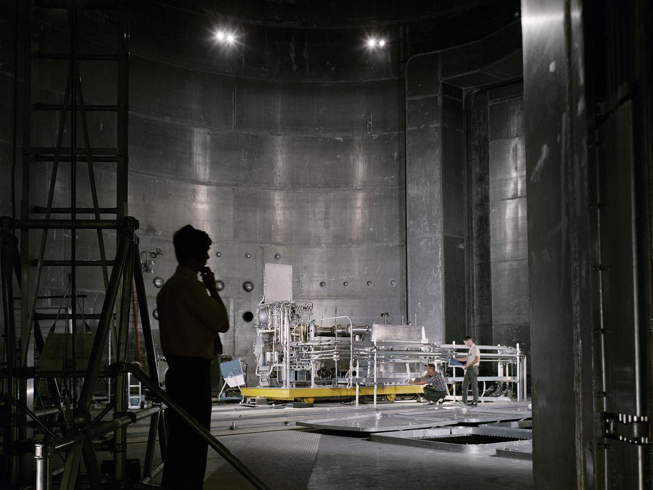 Set up of a Brayton Cycle Power System test in the Space Power Facility’s massive vacuum chamber at the National Aeronautics and Space Administration’s (NASA) Plum Brook Station in Sandusky, Ohio. The $28.4-million facility, which began operations in 1969, is the largest high vacuum chamber ever built. The chamber is 100 feet in diameter and 120 feet high. It can produce a vacuum deep enough to simulate the conditions at 300 miles altitude.  The Space Power Facility was originally designed to test nuclear-power sources for spacecraft, but it was never used for that purpose.   The Space Power Facility was first used to test a 15 to 20-kilowatt Brayton Cycle Power System for space applications. Three different methods of simulating solar heat were employed during the tests.    Lewis researchers studied the Brayton power system extensively in the 1960s and 1970s. The Brayton engine converted solar thermal energy into electrical power. The system operated on a closed-loop Brayton thermodynamic cycle with a helium-xenon gas mixture as its working fluid. A space radiator was designed to serve as the system’s waste heat rejecter. The radiator was later installed in the vacuum chamber and tested in a simulated space environment to determine its effect on the power conversion system. The Brayton system was subjected to simulated orbits with 62 minutes of sun and 34 minutes of shade.