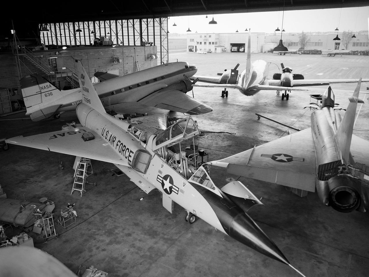 Several aircraft parked inside the Flight Research Building, or hangar, at the National Aeronautics and Space Administration (NASA) Lewis Research Center in Cleveland, Ohio. A Convair F-106B Delta Dart is in the foreground, a Convair F-102A Delta Dagger is to the right, a Douglas DC-3 is in the back to left, and a Convair T-29 is in background. Lewis’ Martin B-57B Canberra is not seen in this photograph. The F-102A had just been acquired by Lewis to serve as a chase plane for the F-106B. The Lewis team removed the weapons system and 700 pounds of wire from the F-106B when it was acquired on October 20, 1966. The staff cut holes in the wings and modified the elevons to mount the test nacelles. A 228-gallon fuel tank was installed in the missile bay, and the existing wing tanks were used for instrumentation. This photograph contains a rare view of the Block House, seen to the left of the aircraft. Lewis acquired three large developmental programs in 1962—the Centaur and Agena rockets and the M-1 engine. The center was short on office space at the time, and its flight research program was temporarily on the wane. Lewis management decided to construct a large cinderblock structure inside one half of the hangar to house the new personnel. This structure was used until 1965 when the new Developmental Engineering Building was built. The Block House was eventually torn down in 1973.