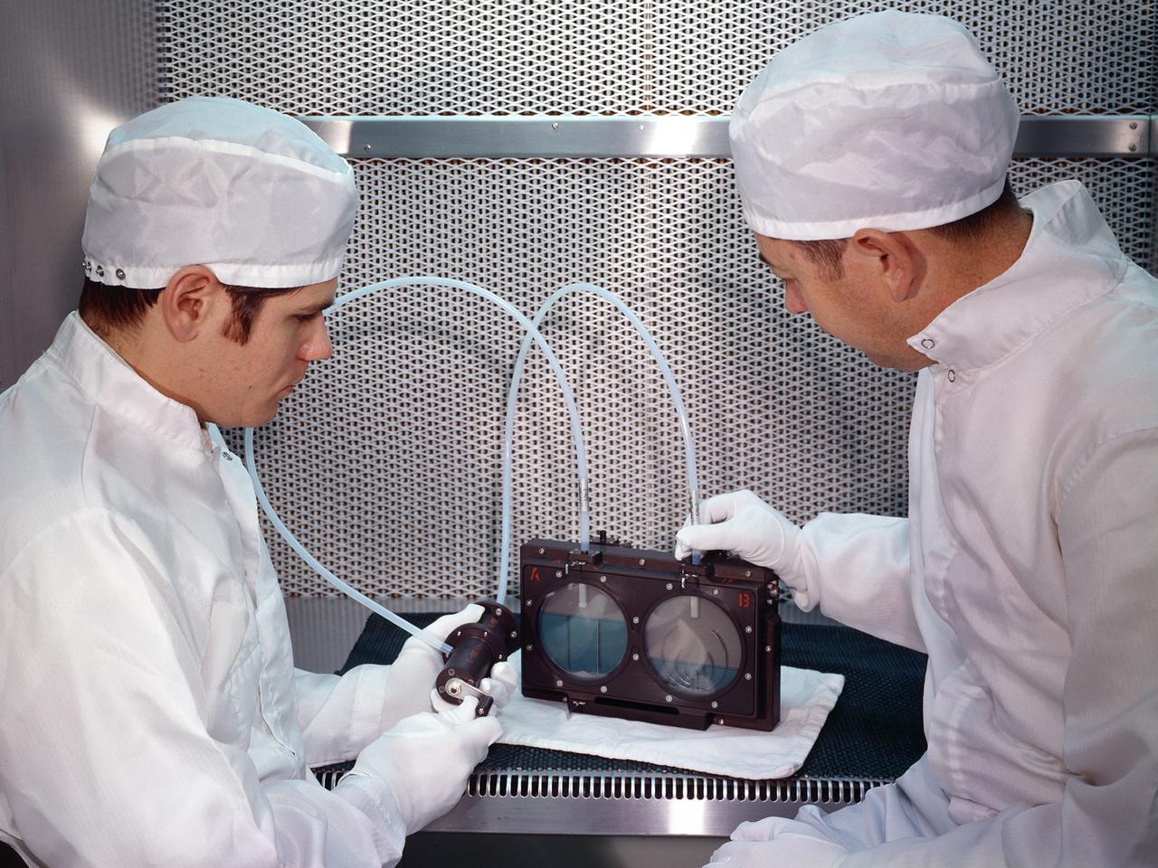 Two researchers at the National Aeronautics and Space Administration (NASA) Lewis Research Center demonstrate the test equipment they devised to study the transfer of liquid in microgravity onboard the Apollo 14 mission. The test was an early step in developing the ability to transfer liquids from a tanker vehicle to spacecraft in space. Researchers needed to know the tank’s outflow characteristics, the fluid’s behavior when entering new tank, and the effects of accelerations.    Others had performed some calculations and analytical studies, but no one had examined the complete transfer from one tank to another in microgravity. The early calculations concluded that the transfer process was impossible without devices to control the liquid and gas.     This investigation specifically sought to demonstrate the effectiveness of two different surface-tension baffle designs. The experiment was an entirely closed system with two baffled-tanks. The researchers also built a similar device without the baffles. The experiment was carried onboard the Apollo 14 spacecraft and conducted during the coast period on the way to the moon. The two surface tension baffle designs in the separate tanks were shown to be effective both as supply tanks and as receiver tanks. The liquid transferred within two percent of the design value with ingesting gas. The unbaffled tanks ingested gas after only 12-percent of the fluid had transferred.