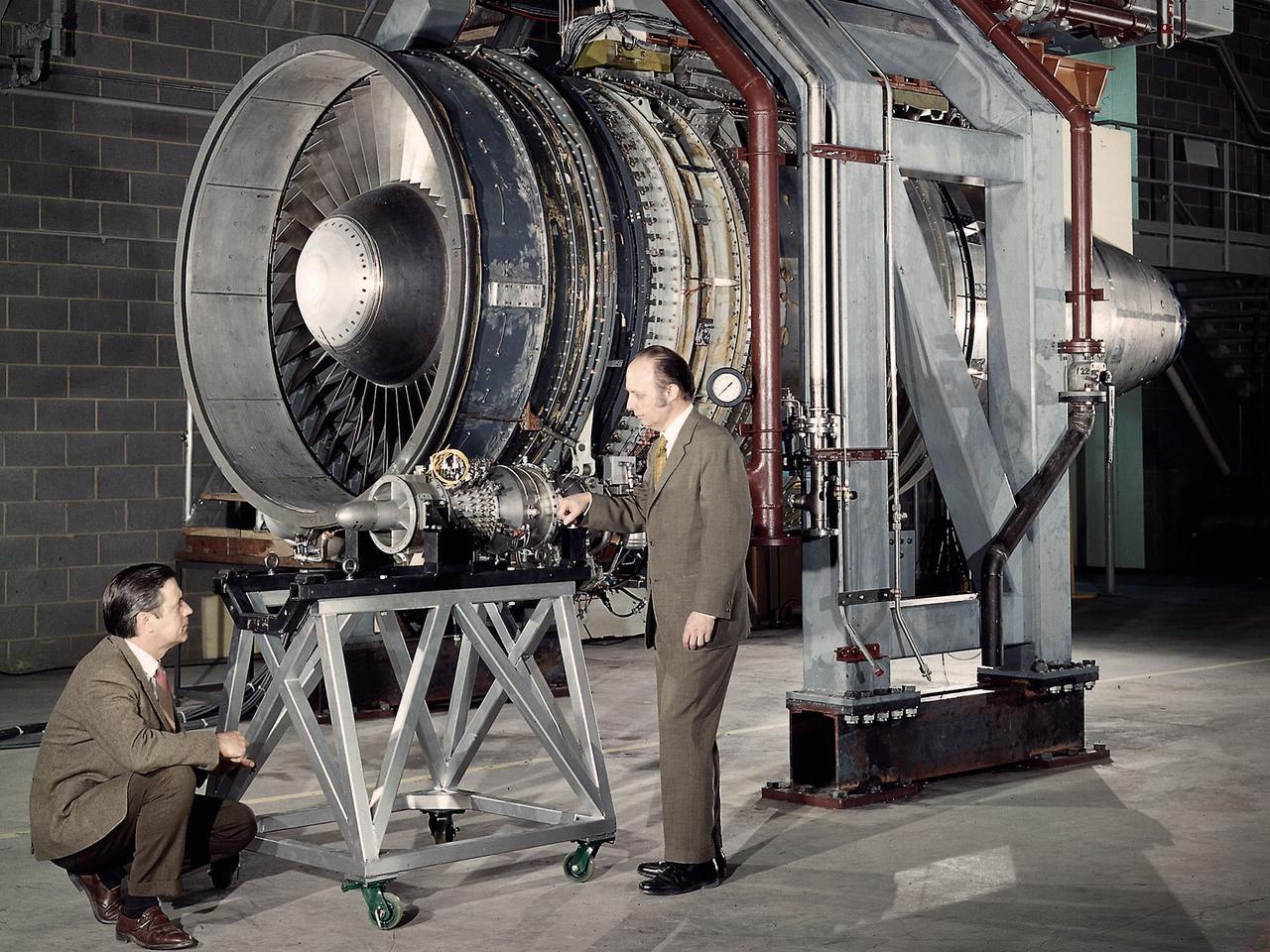 Researchers Robert Cummings, left, and Harold Gold with the small Low Cost Engine in the shadow of the much larger Quiet Engine at the National Aeronautics and Space Administration (NASA) Lewis Research Center. The two engines were being studied in different test cells at the Propulsion Systems Laboratory. Jet engines had proven themselves on military and large transport aircraft, but their use on small general aviation aircraft was precluded by cost. Lewis undertook a multiyear effort to develop a less expensive engine to fill this niche using existing technologies.  Lewis researchers designed a four-stage, axial-flow engine constructed from sheet metal. It was only 11.5 inches in diameter and weighed 100 pounds. The final design specifications were turned over to a manufacturer in 1972. Four engines were created, and, as expected, the fabrication and assembly of the engine were comparatively inexpensive. In 1973 the Low Cost Engine had its first realistic analysis in the Propulsion Systems Laboratory altitude tank. The engine successfully operated at speeds up to Mach 1.24 and simulated altitudes of 30,000 feet.    NASA released the engine to private industry in the hope that design elements would be incorporated into future projects and reduce the overall cost of small jet aircraft. Small jet and turboprop engines became relatively common in general aviation aircraft by the late 1970s.