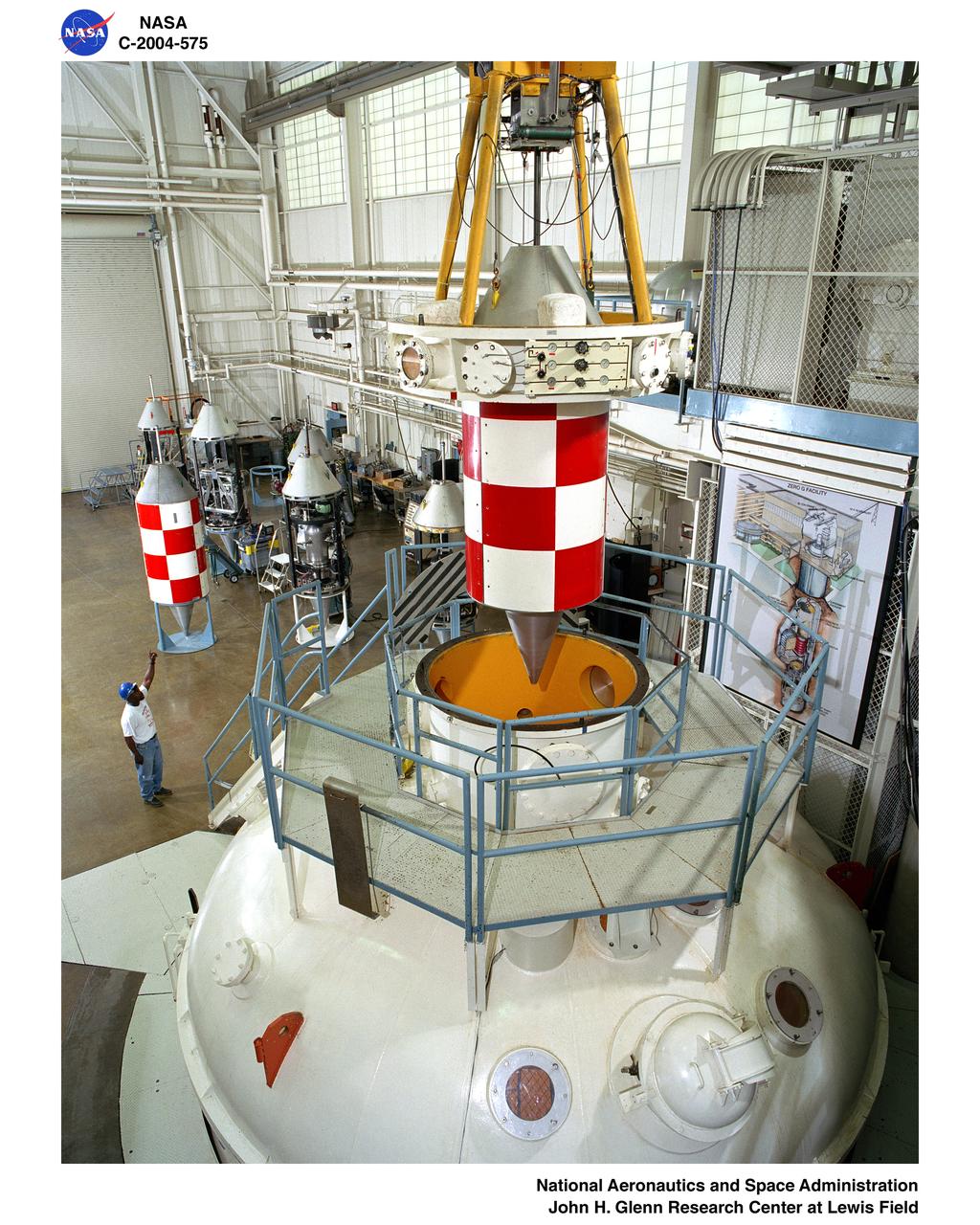 Mezzanine view of the drop vehicle and release mechanism being positioned over the vacuum chamber with a technician signaling the crane operator in the Zero Gravity Research Facility.