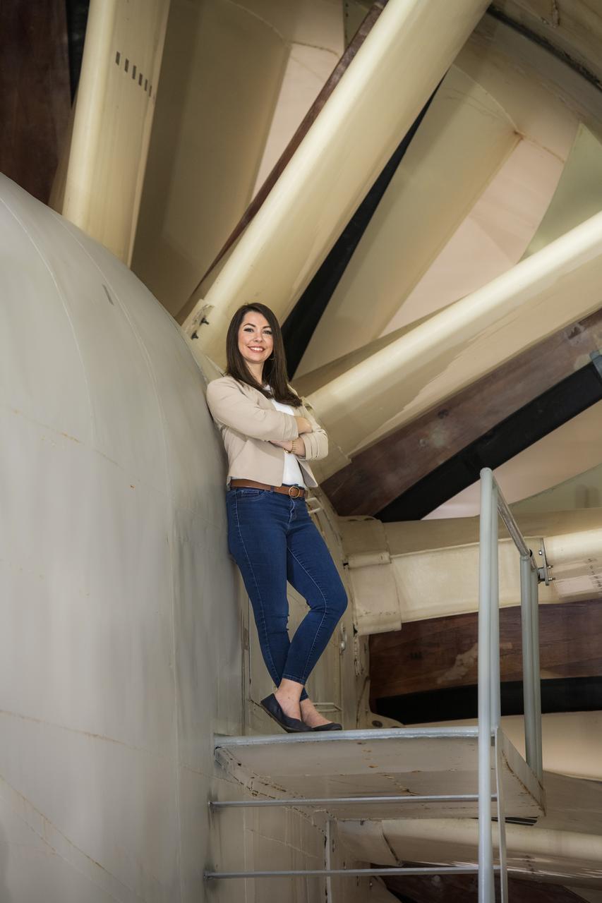 Emily Timko, featured in a Faces of NASA article, poses in the IRT (Icing Research Tunnel) where she works as a “cloud engineer”.  She is a Mechanical Test Engineer and works to create unique water spray conditions that simulate icing clouds in the natural aircraft flight environment.  Shown in the photo is a portion of the fan drive motor and fan blades that together drive the air through the wind tunnel.