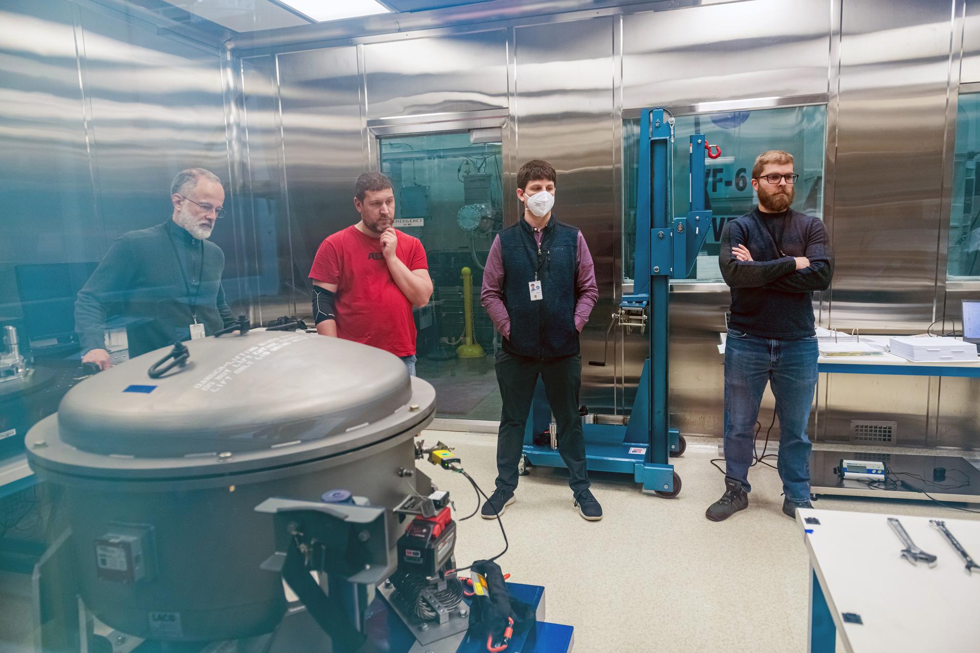 Four men stand inside a clean, metallic room at NASA Glenn Research Center, next to a large cylindrical component secured to a platform in the foreground. One man is wearing a red shirt, another wears a mask, and the others are dressed in dark clothing.