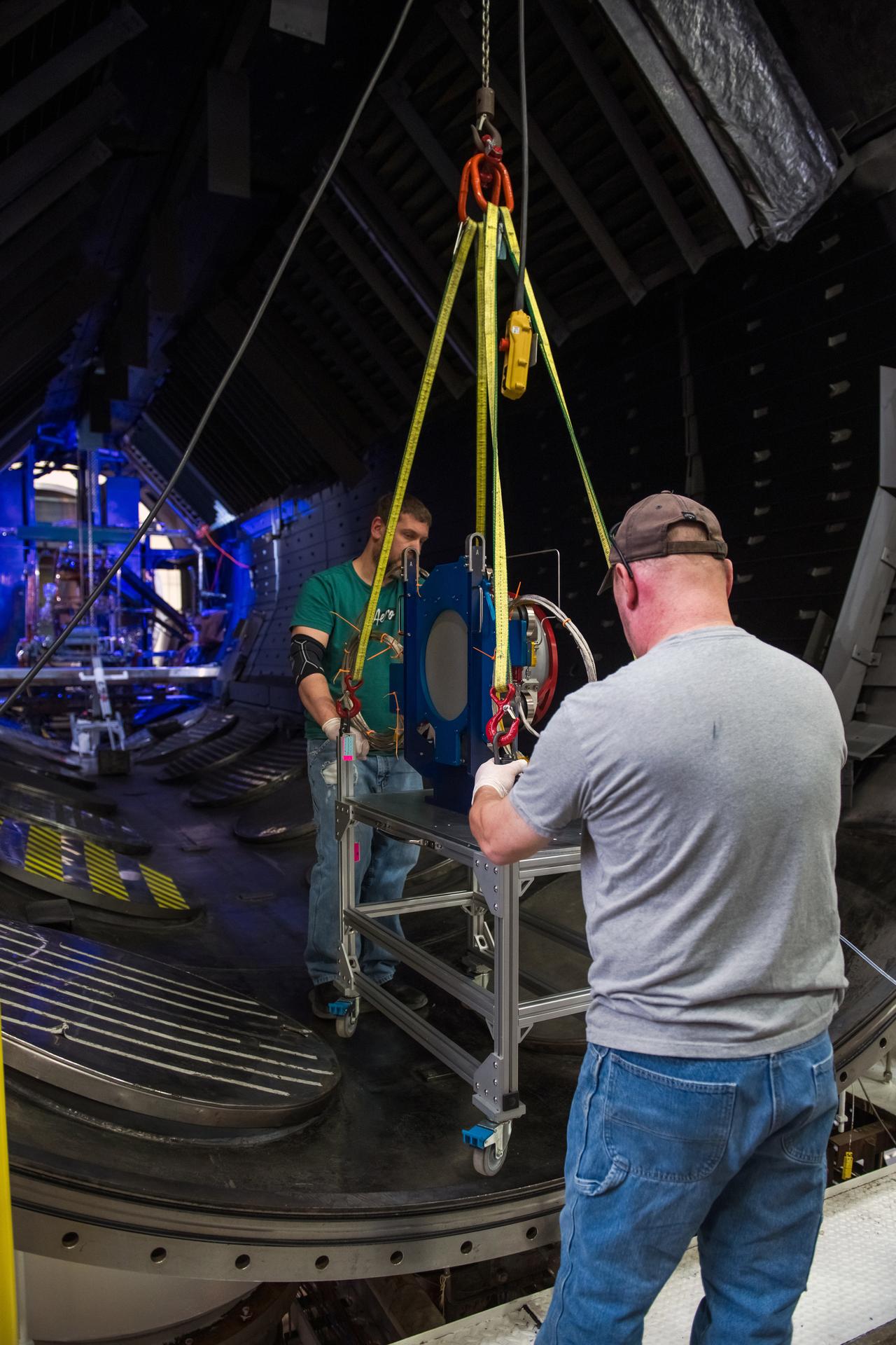 Two technicians guide an Advanced Electric Propulsion System (AEPS) thruster being lowered by a yellow hoist inside a vacuum chamber at NASA Glenn Research Center. The thruster is secured with yellow lifting straps and positioned on a metal cart with wheels. The technician in the foreground wears a gray shirt and cap, while the other stands across in a green shirt. The chamber’s interior features ribbed walls and dark, curved flooring, with the test setup visible in the background.